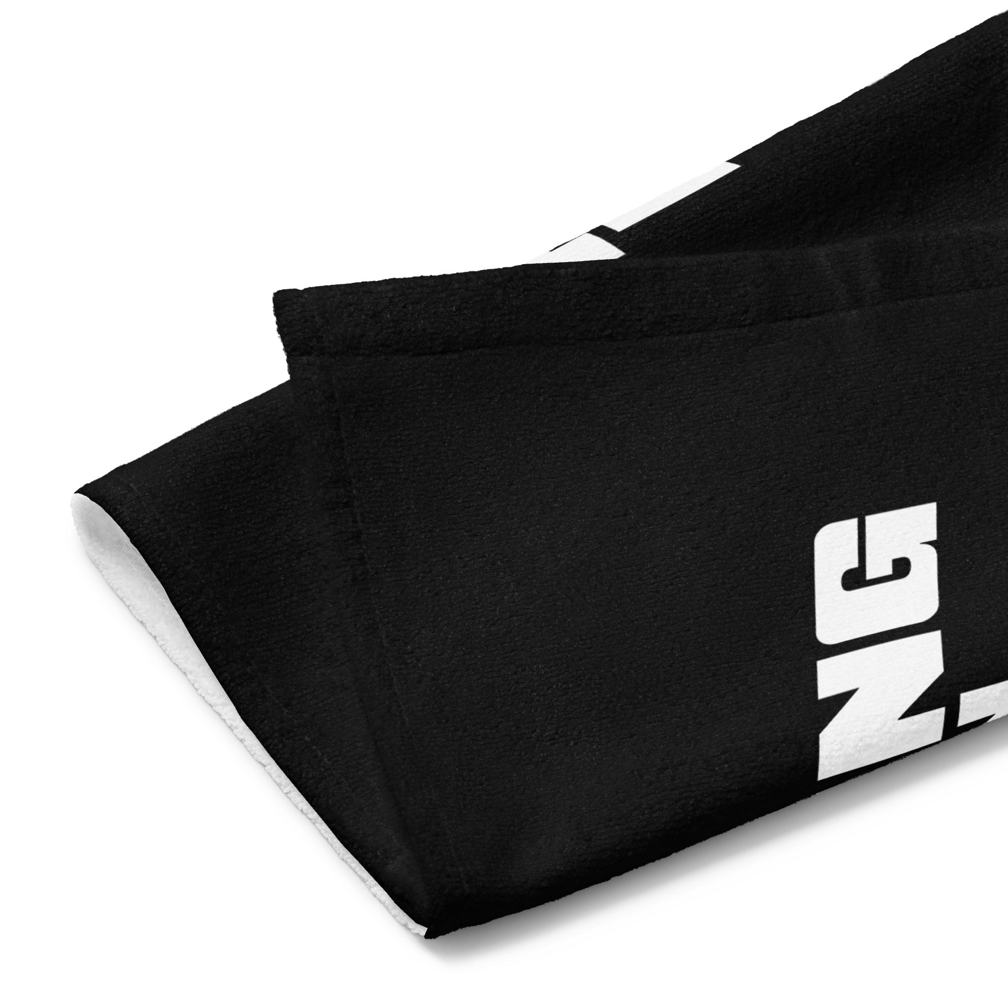 Close-up of the towel corner folded over, revealing the black front with partial white lettering and a white terry cloth backing, showing the soft textured edge and stitching.