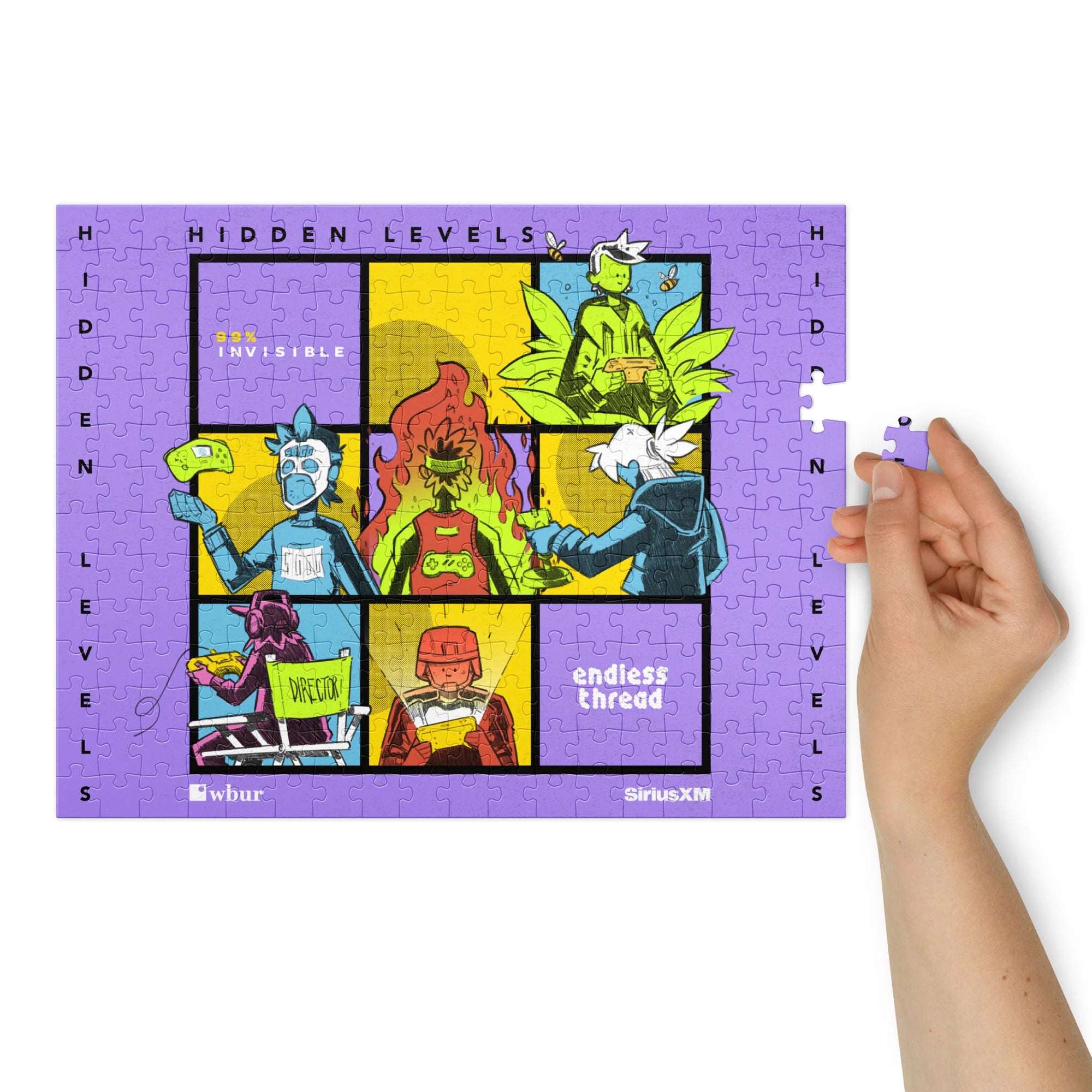 Close-up of a hand holding a puzzle piece near the right edge of a mostly completed rectangular jigsaw puzzle. The design features the same six-panel comic-style artwork with bright characters and text reading 'HIDDEN LEVELS,' '99% Invisible,' and 'Endless Thread,' bordered in purple with 'WBUR' and 'SiriusXM' logos along the bottom.