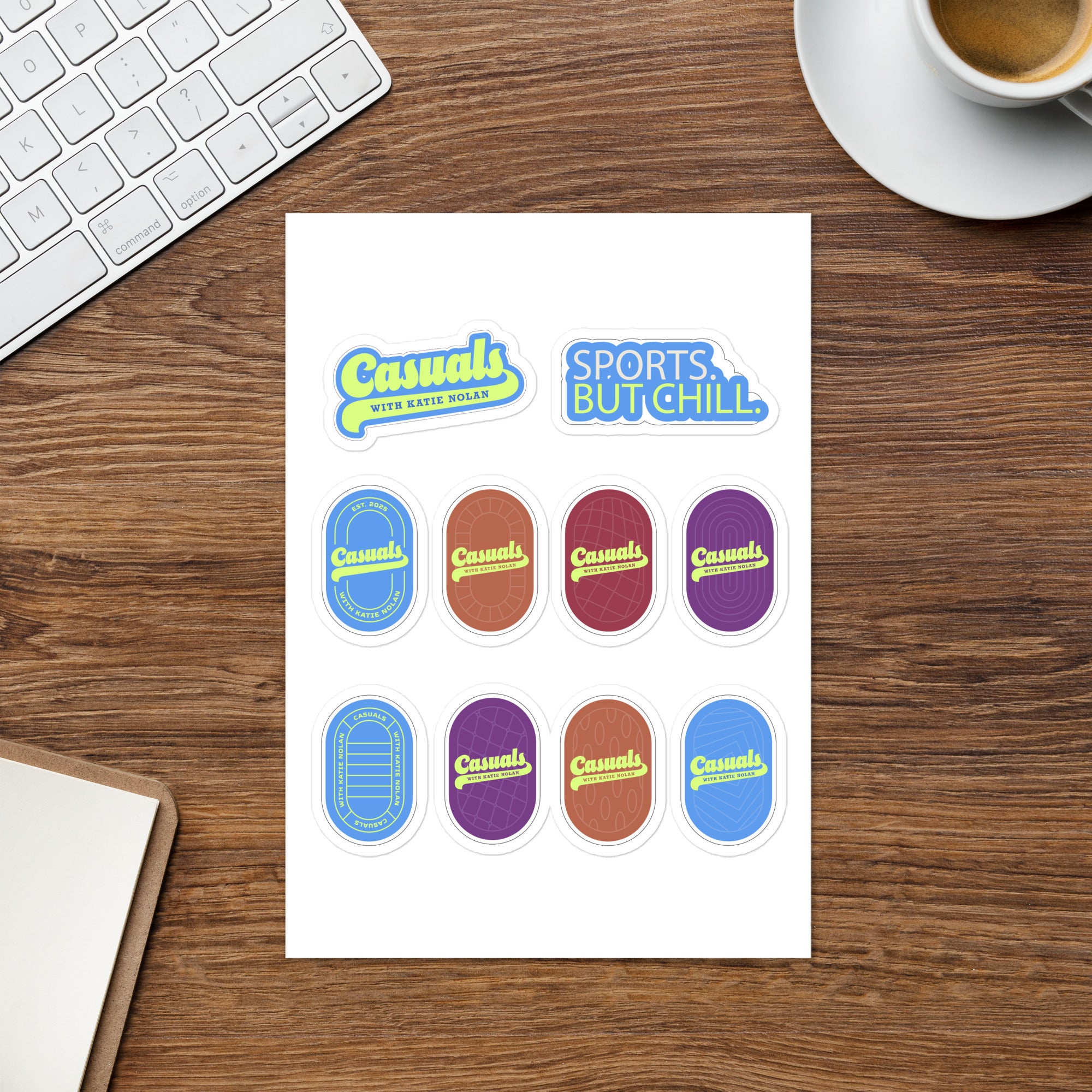 Overhead view of the same white rectangular 'Casuals with Katie Nolan' sticker sheet placed on a wooden desk beside a computer keyboard and a cup of coffee. The sheet displays two text stickers at the top and eight oval logo designs below in blue, red, purple, and brown.