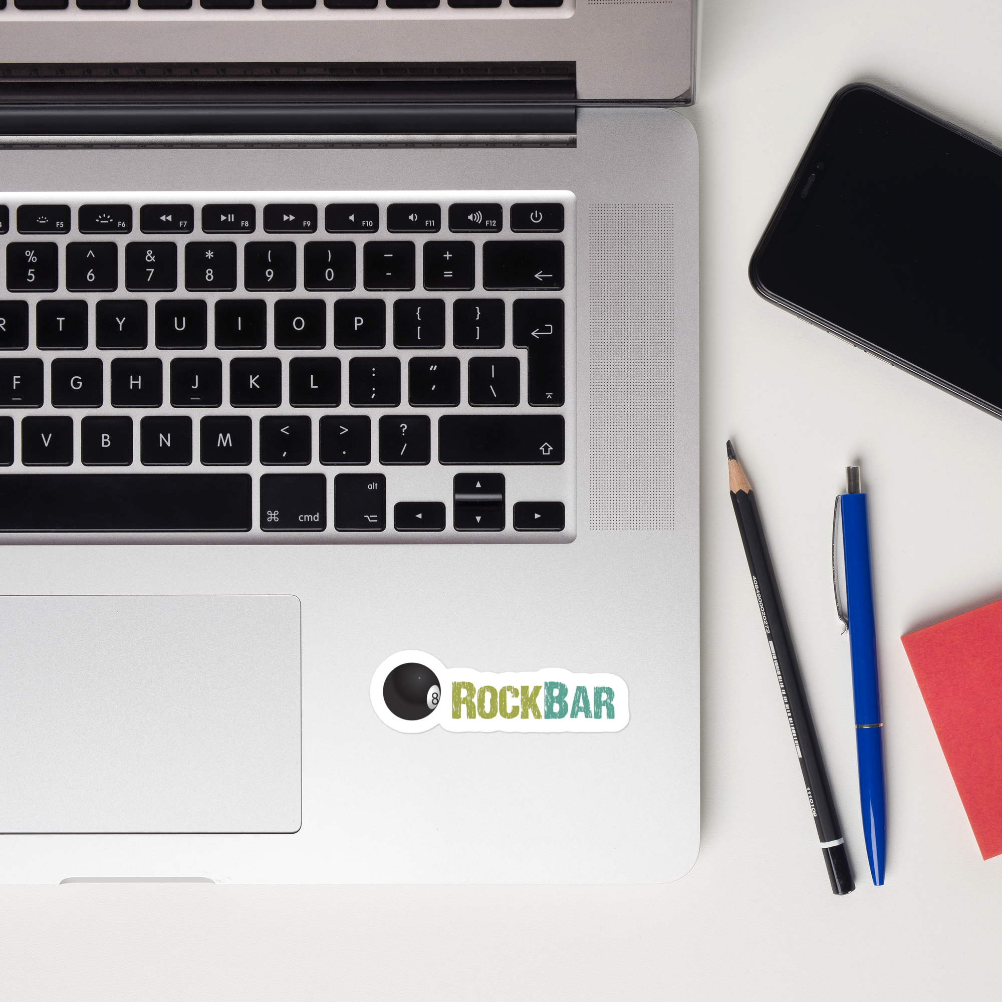 A laptop keyboard with a 'ROCKBAR' sticker, a phone, pens, and a notepad on a white surface.