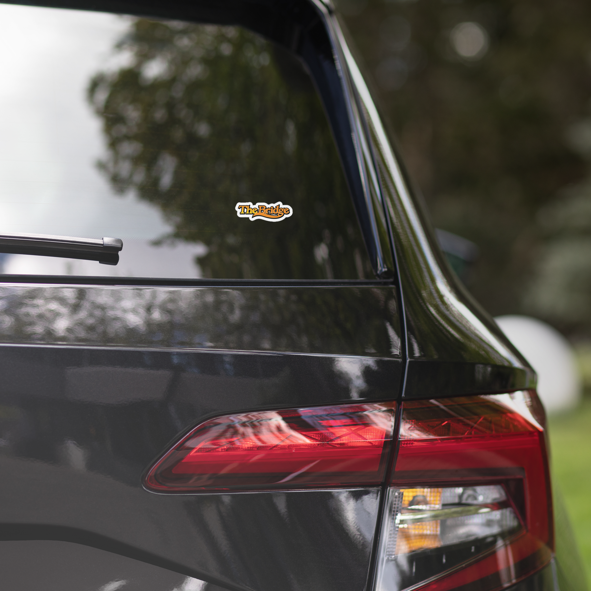 Close-up of a black vehicle's rear, displaying a 'The Bridge' sticker on the window.