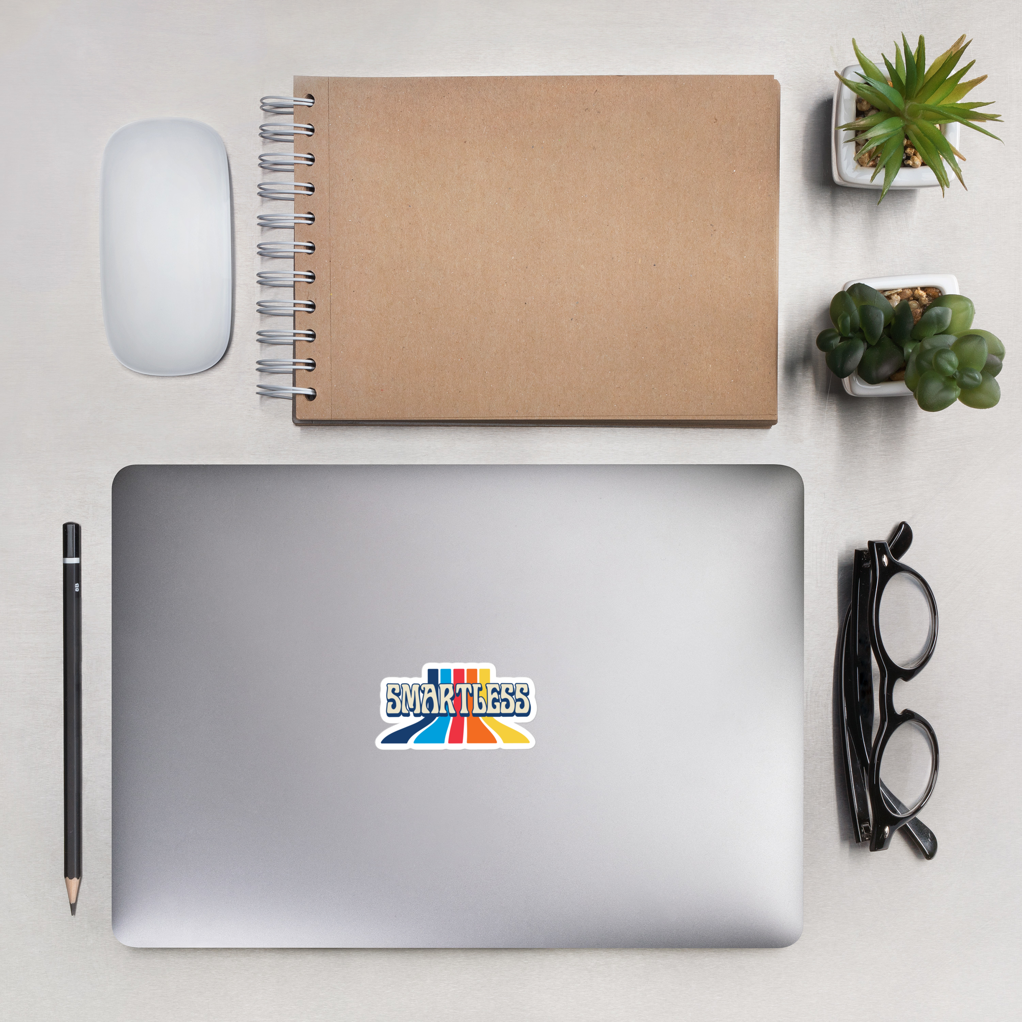 A flat lay of a computer  with a "SmartLess" sticker on a desk surrounded by a mouse, notebook, pen, eye glasses and small potted plants.