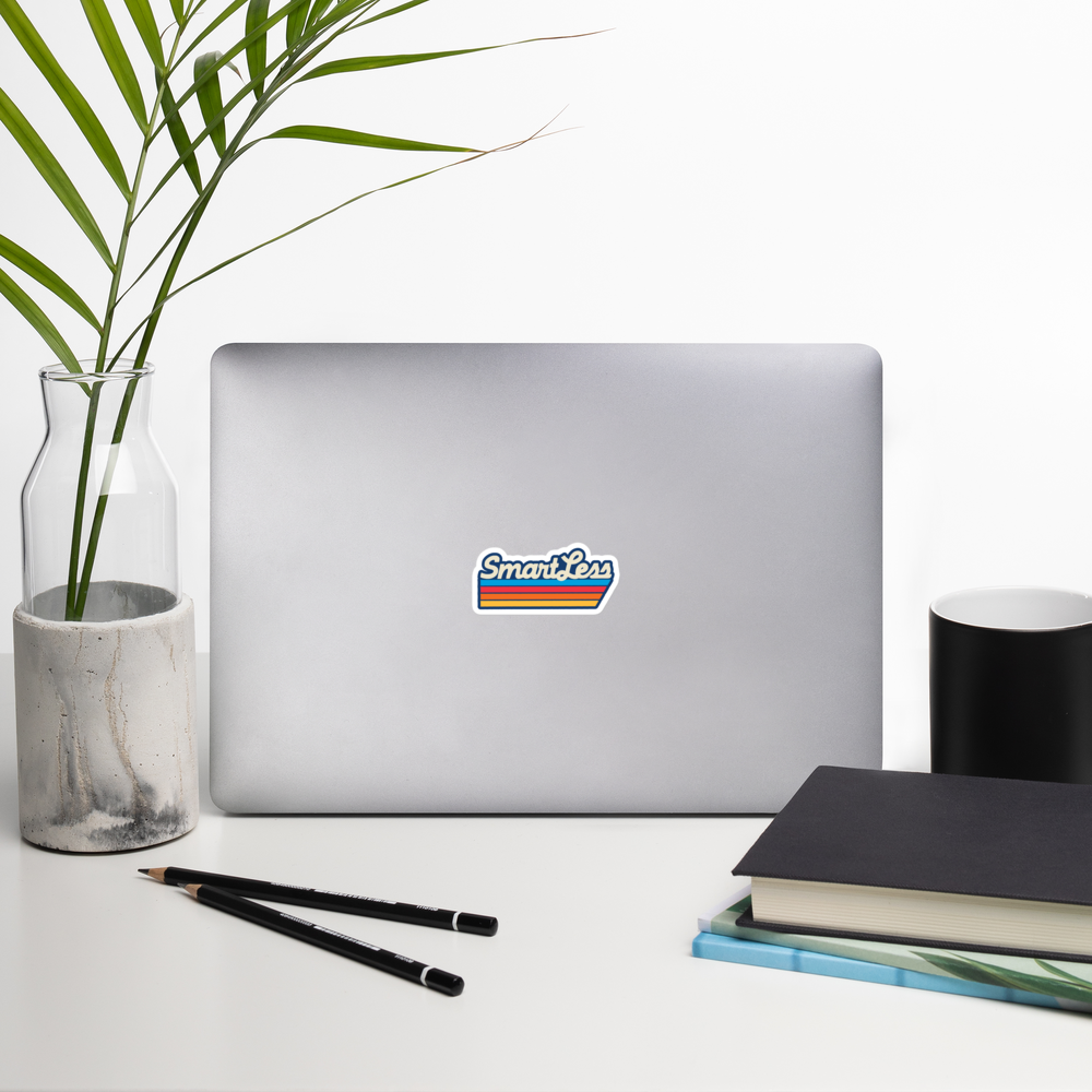 A laptop with a 'SmartLess' sticker on it, surrounded by a plant in a vase, a black mug, notebooks, and pencils on a white desk.
