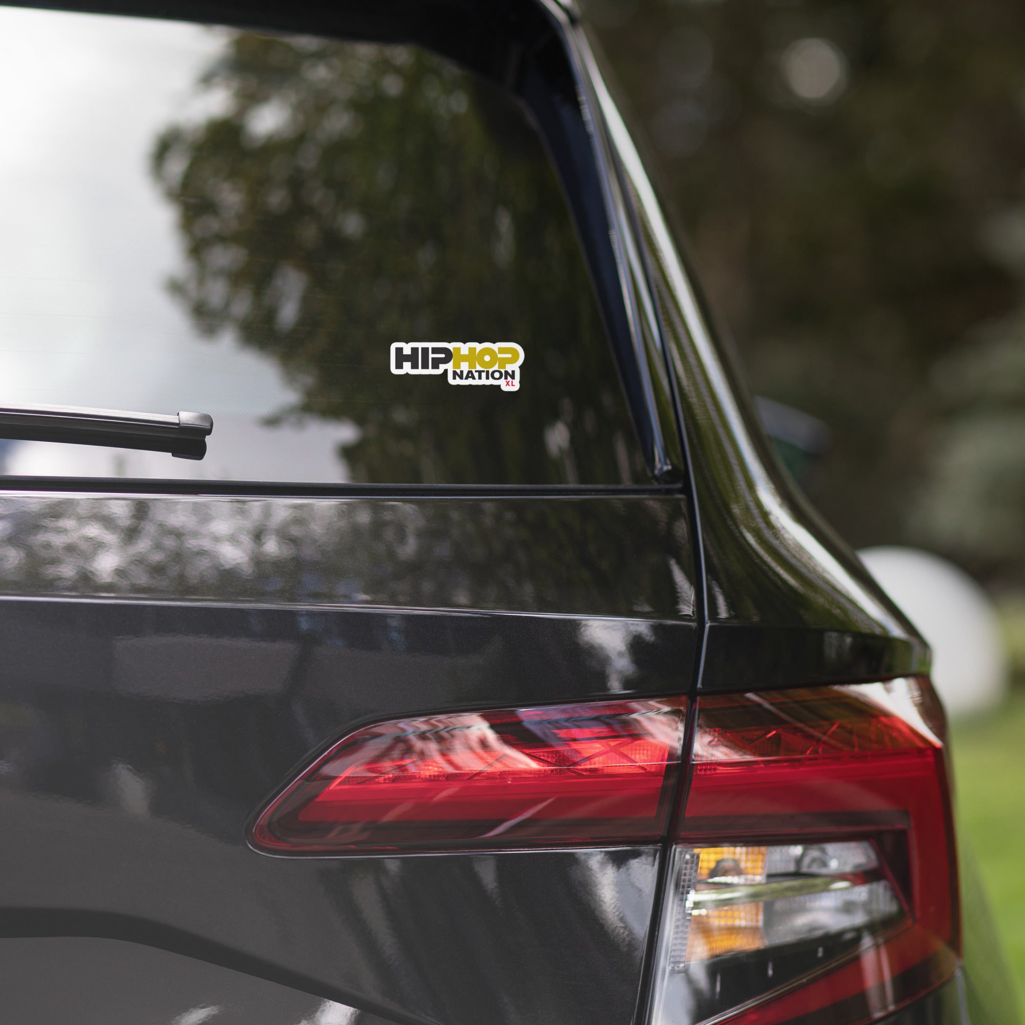 Close-up of a black car's rear with a 'HIP HOP NATION' sticker in black, white, and gold on the window and a visible tail light.