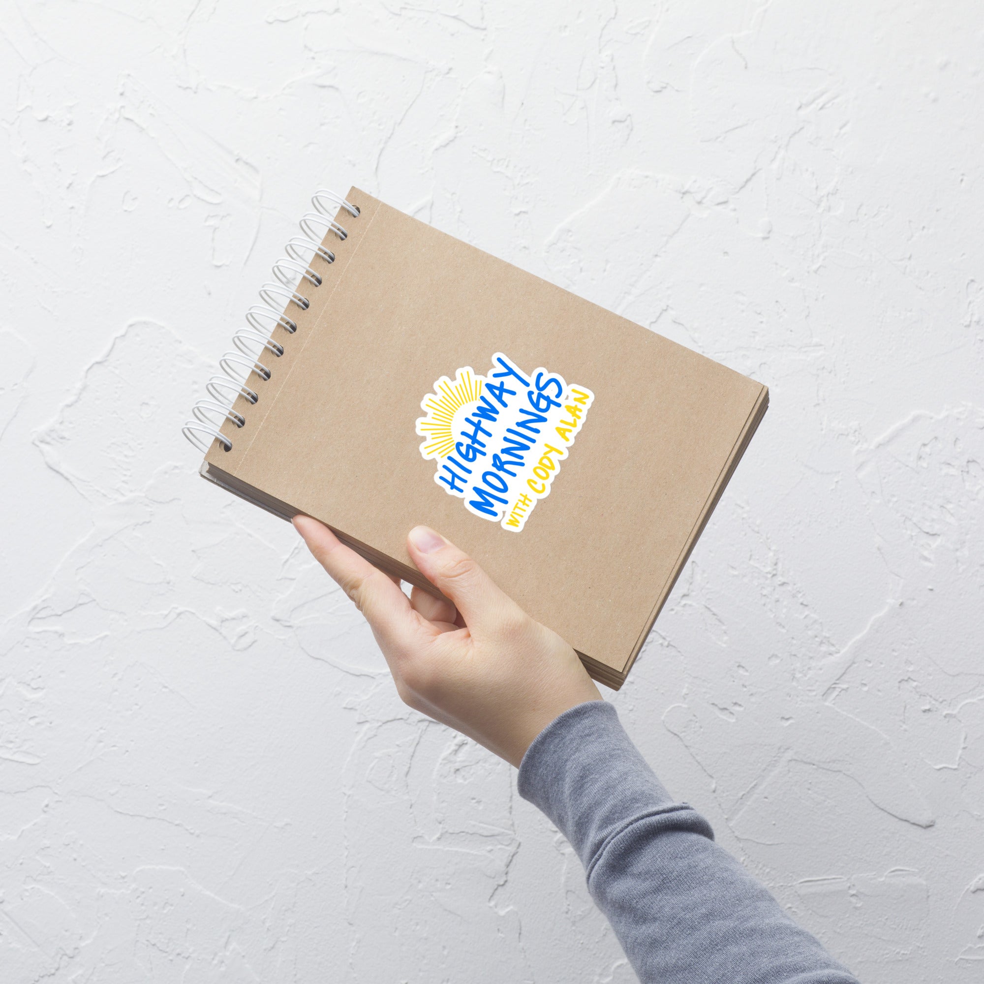 A hand holding a brown spiral-bound notebook with the 'HIGHWAY MORNINGS WITH CODY ALAN' kiss-cut sticker adhered to the cover, showing the blue and yellow text with a yellow sunburst design.