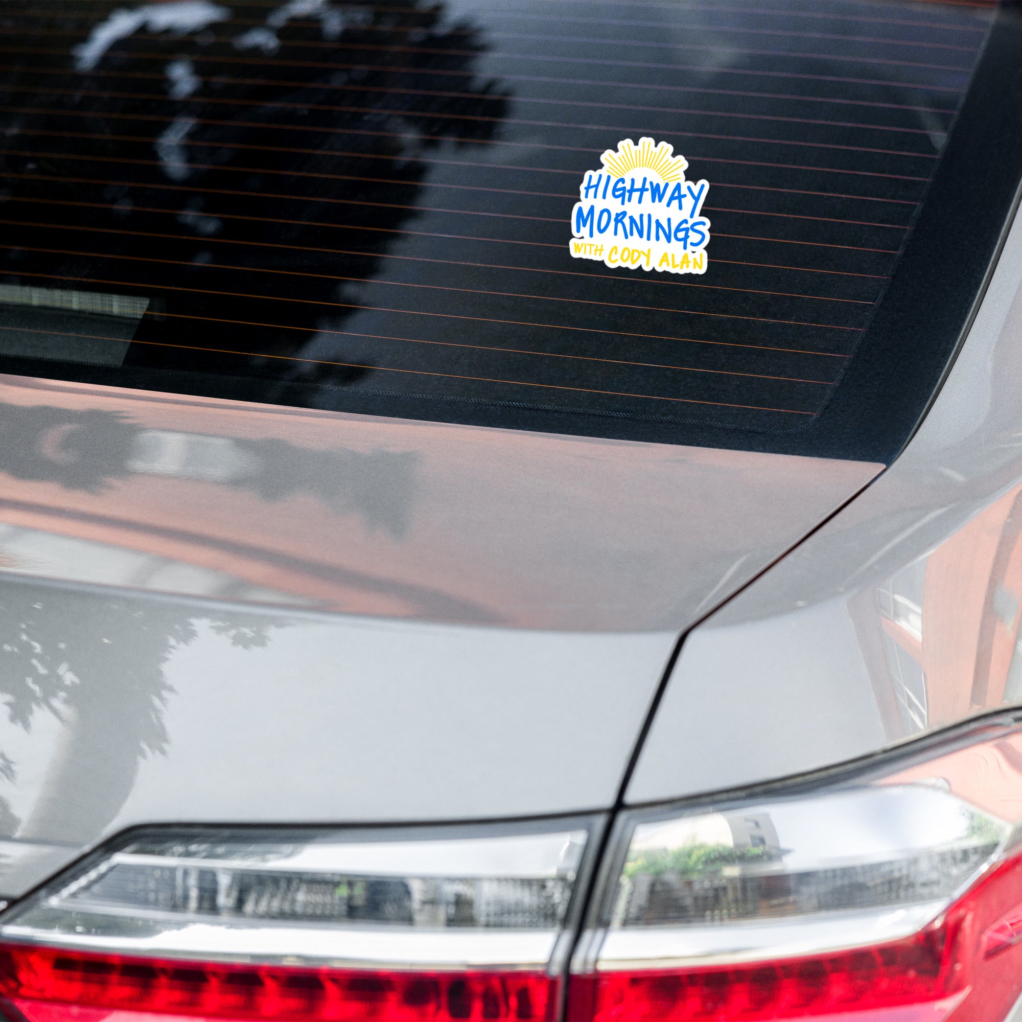 A 'HIGHWAY MORNINGS WITH CODY ALAN' sticker applied to the rear windshield of a silver car, displaying blue and yellow text with a yellow sunburst above.