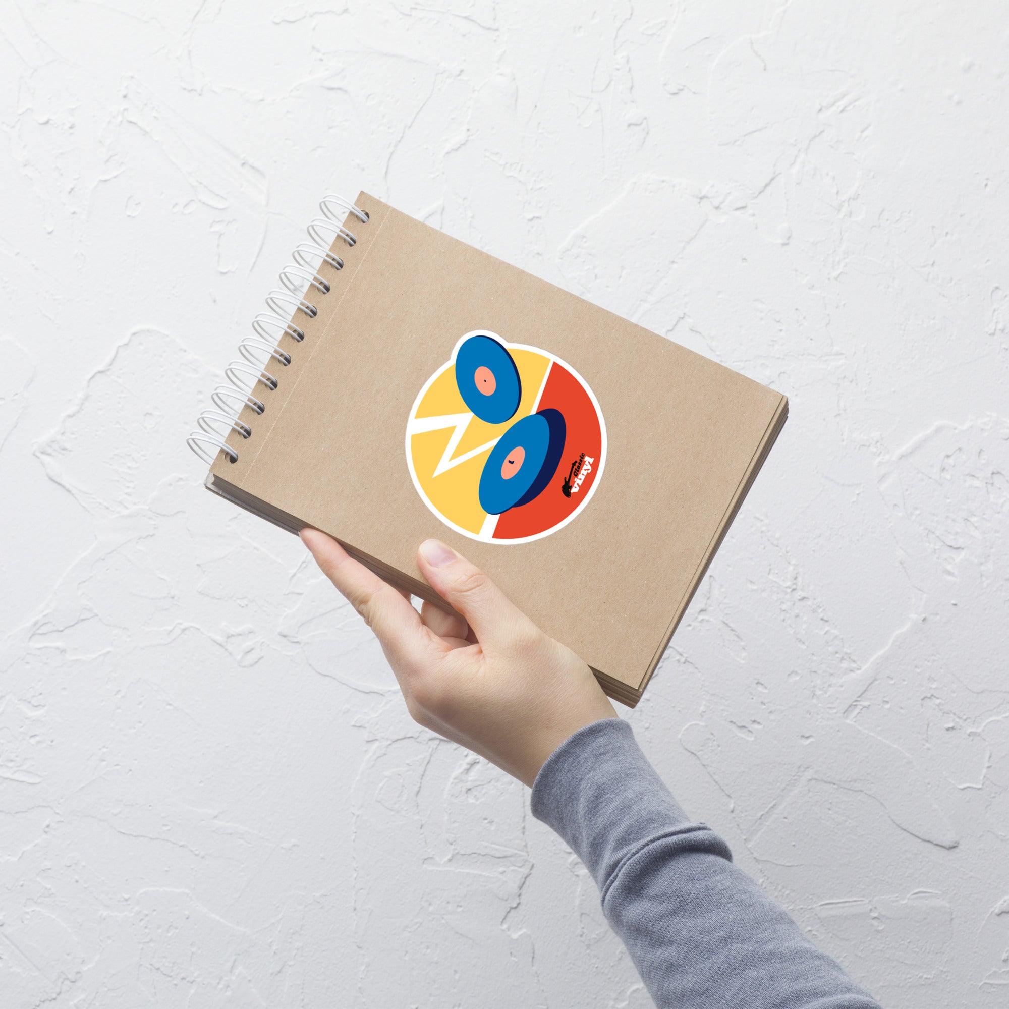A hand holding a spiral notebook with a brown kraft cover, shown against a white textured wall. A circular sticker on the cover features two blue vinyl records on a red and yellow background with a white zigzag lightning shape and the 'Classic vinyl' logo at the bottom.