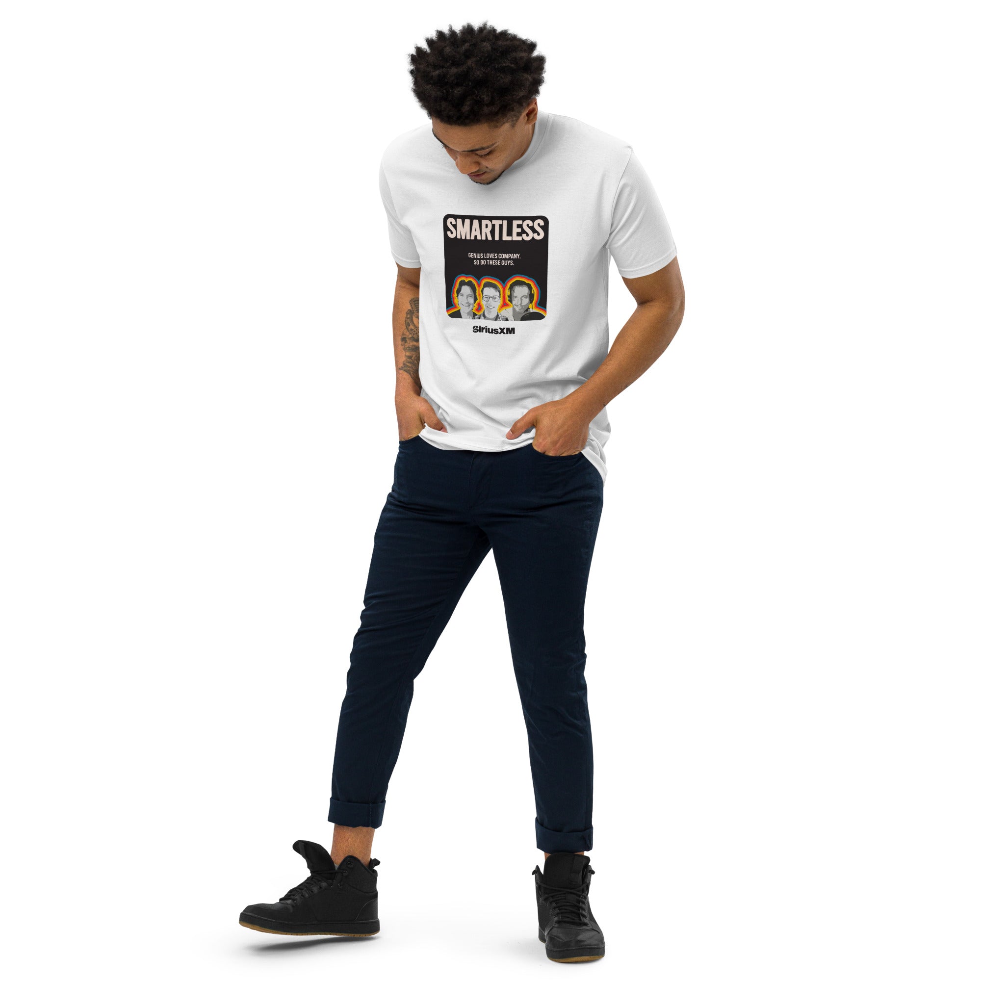 A man wearing a white T-shirt with the 'SMARTLESS' podcast graphic on the front, standing with hands in his pockets and looking down. He pairs the shirt with dark blue pants and black high-top sneakers.