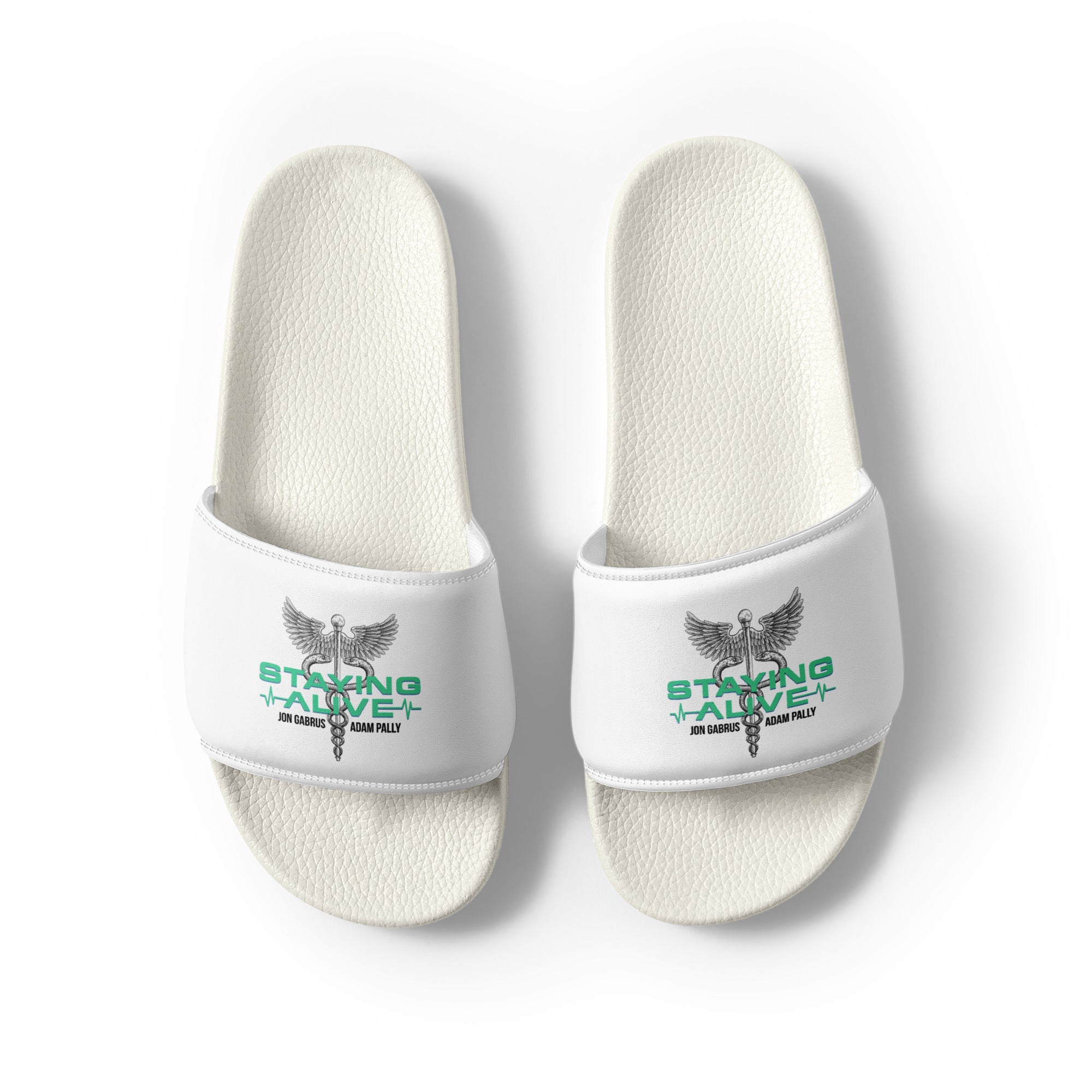 Top-down view of white slide sandals. Each strap features a winged staff with two snakes (caduceus), green text 'STAYING ALIVE', and black text 'JON GABRUS' and 'ADAM PALLY'.