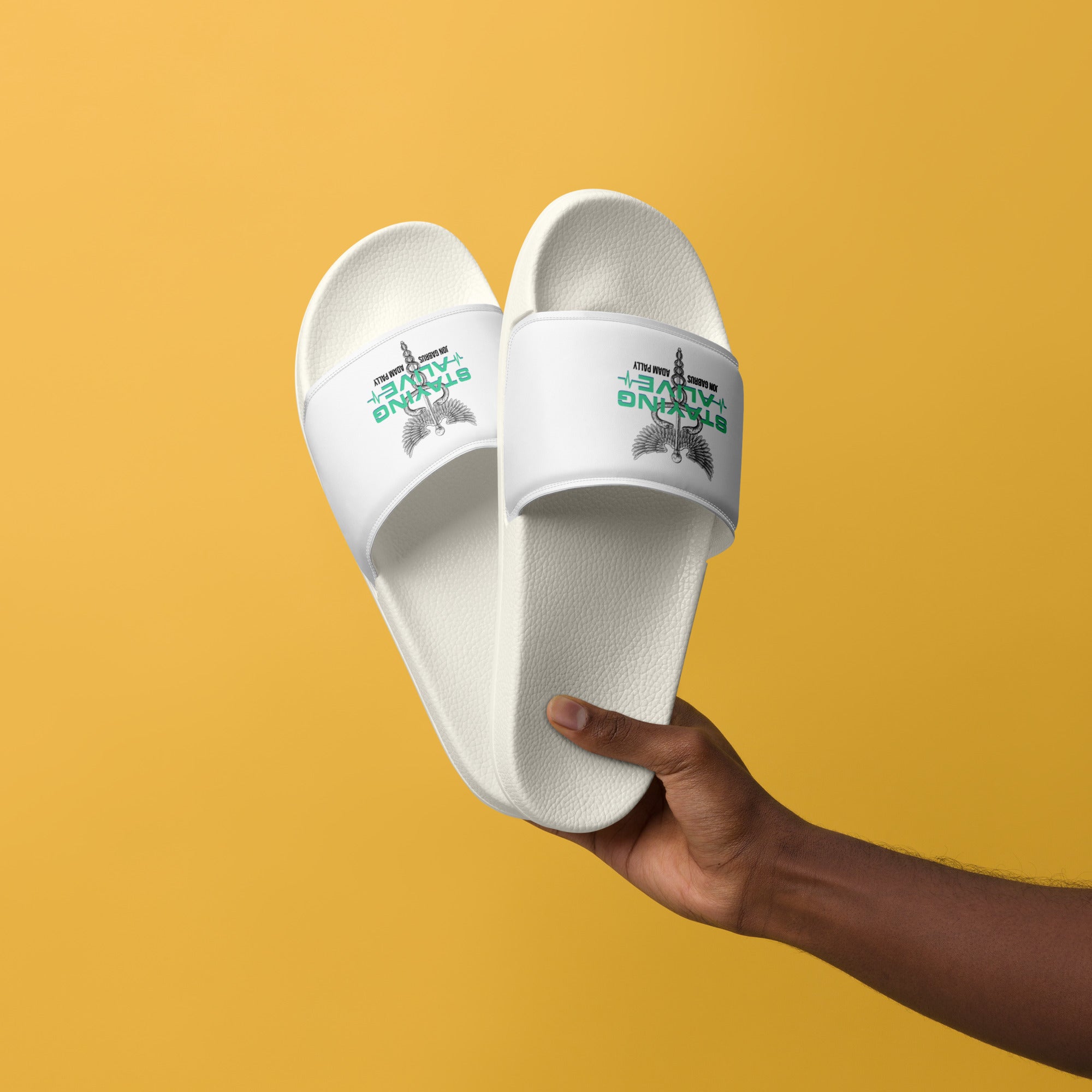 Hand holding white slide sandals in front of a yellow background. Straps show a caduceus (winged staff with two snakes), 'STAYING ALIVE' in green, and names below.