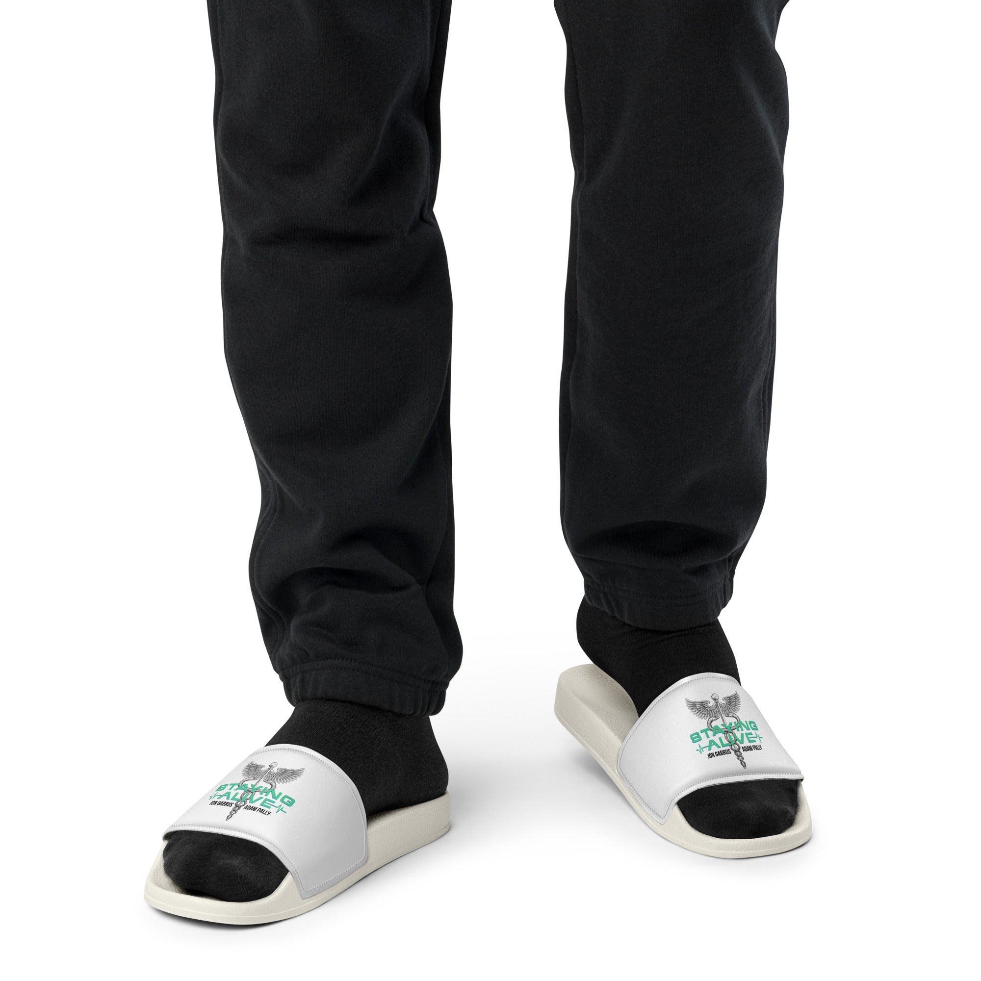 Person wearing white slide sandals with black socks. The straps display a caduceus symbol (winged staff with two snakes) above green 'STAYING ALIVE' text and the host's names in black.