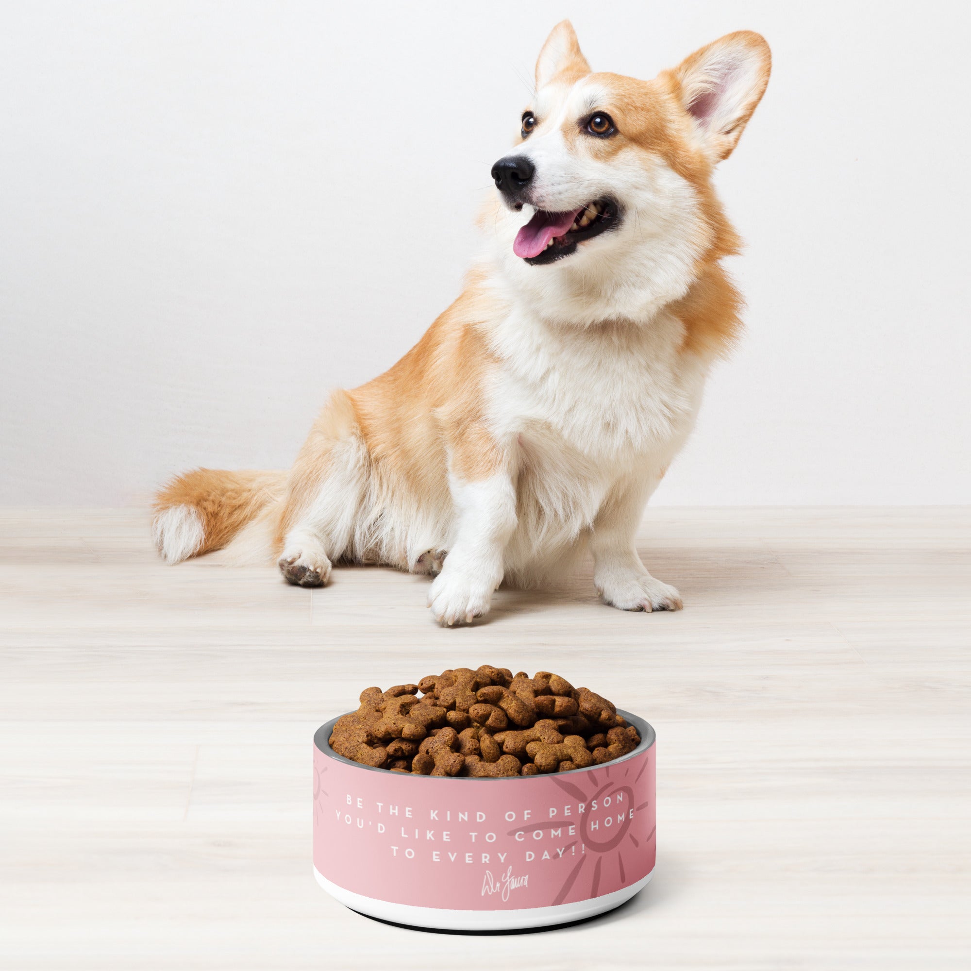 A happy corgi sits beside a pink bowl of dog food with an inspirational 'Dr. Laura' quote on it.