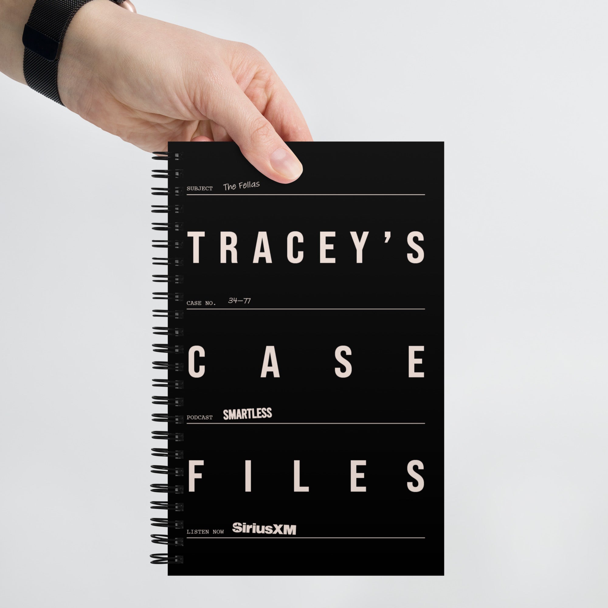 A hand holds a black spiral notebook vertically. The cover resembles a classified case file with the title ‘TRACEY’S CASE FILES’ in bold white text across three sections, and includes fields like ‘Subject: The Fellas’, ‘Case No.: 34–77’, and ‘Podcast: SmartLess’. The bottom line reads ‘Listen Now: SiriusXM’.