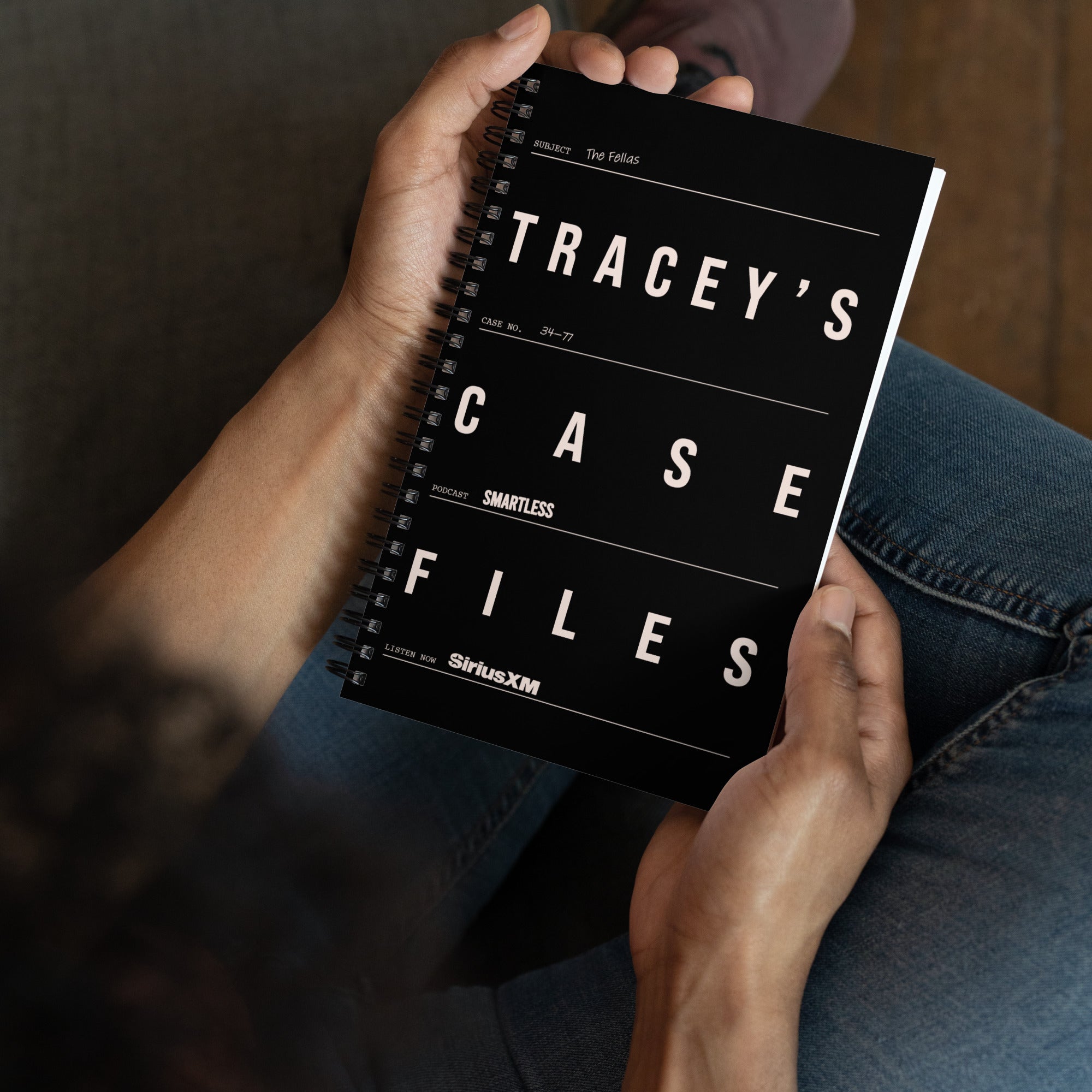 Person sitting and holding the black spiral notebook in their lap. The cover is styled like an evidence file with the title ‘TRACEY’S CASE FILES’, including details like case number, subject, and 'SiriusXM' and 'SmartLess' logos.