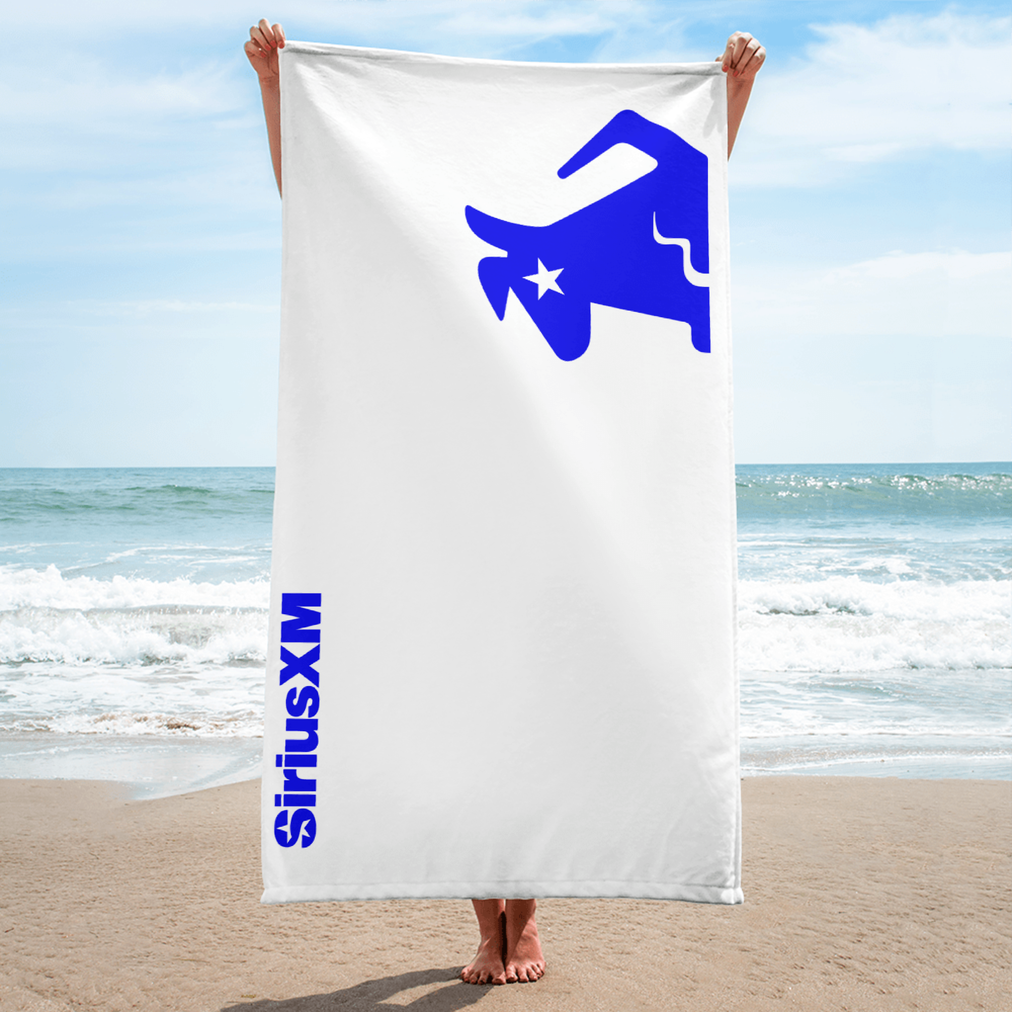 Person at the beach holding a large white towel vertically, featuring a blue 'SiriusXM' logo and Stella dog design.