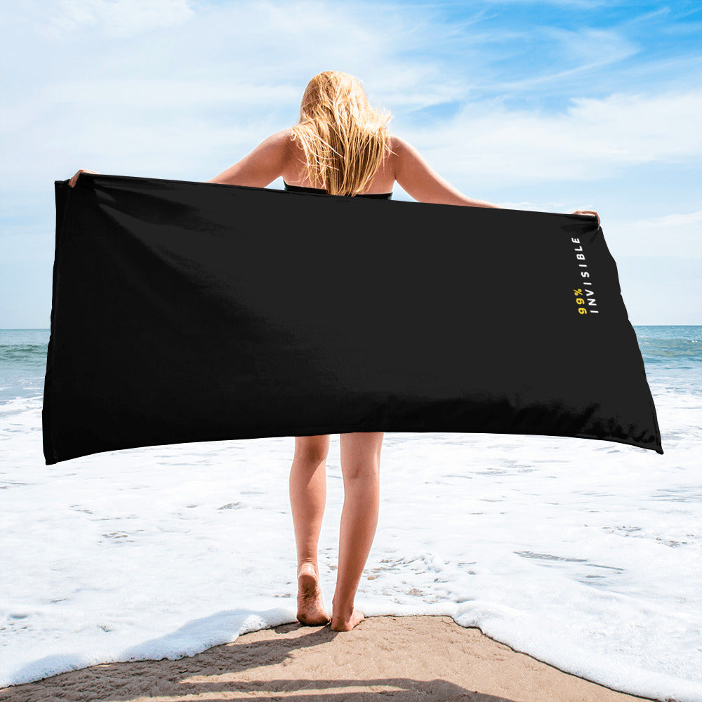 A woman standing on the beach facing the ocean, holding a large black towel that reads '99% INVISIBLE' in white and yellow text in the bottom right corner.