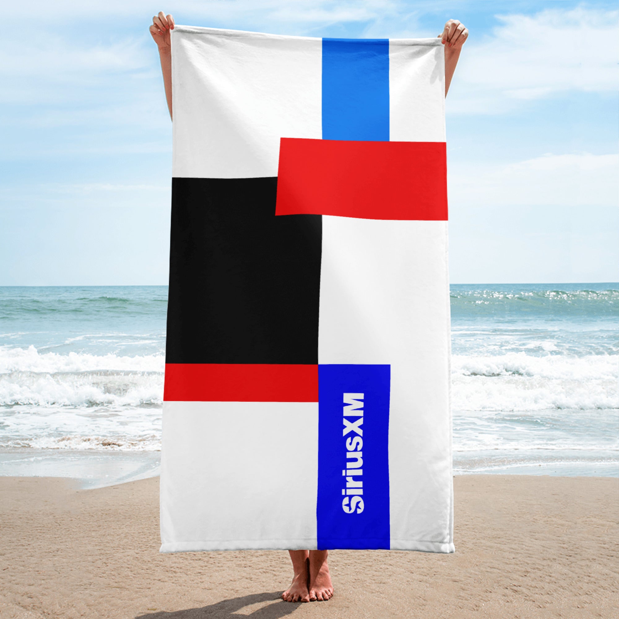 A person standing on the beach holding a vertically oriented towel with a modern geometric design in red, blue, black, and white, displaying the 'SiriusXM' logo in white on a blue vertical strip near the bottom.