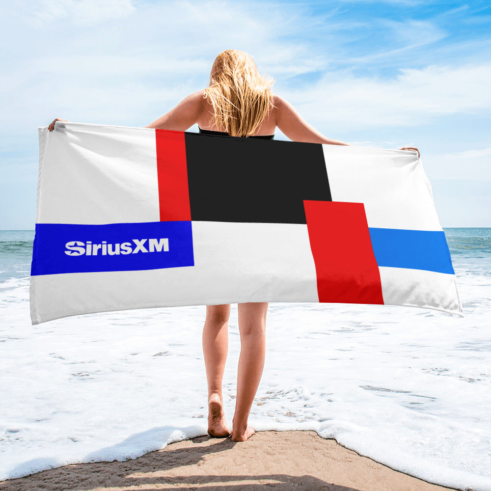 A woman standing on the beach holding a large towel with a bold geometric design in red, blue, black, and white, featuring the 'SiriusXM' logo in white on a blue block near the left side.