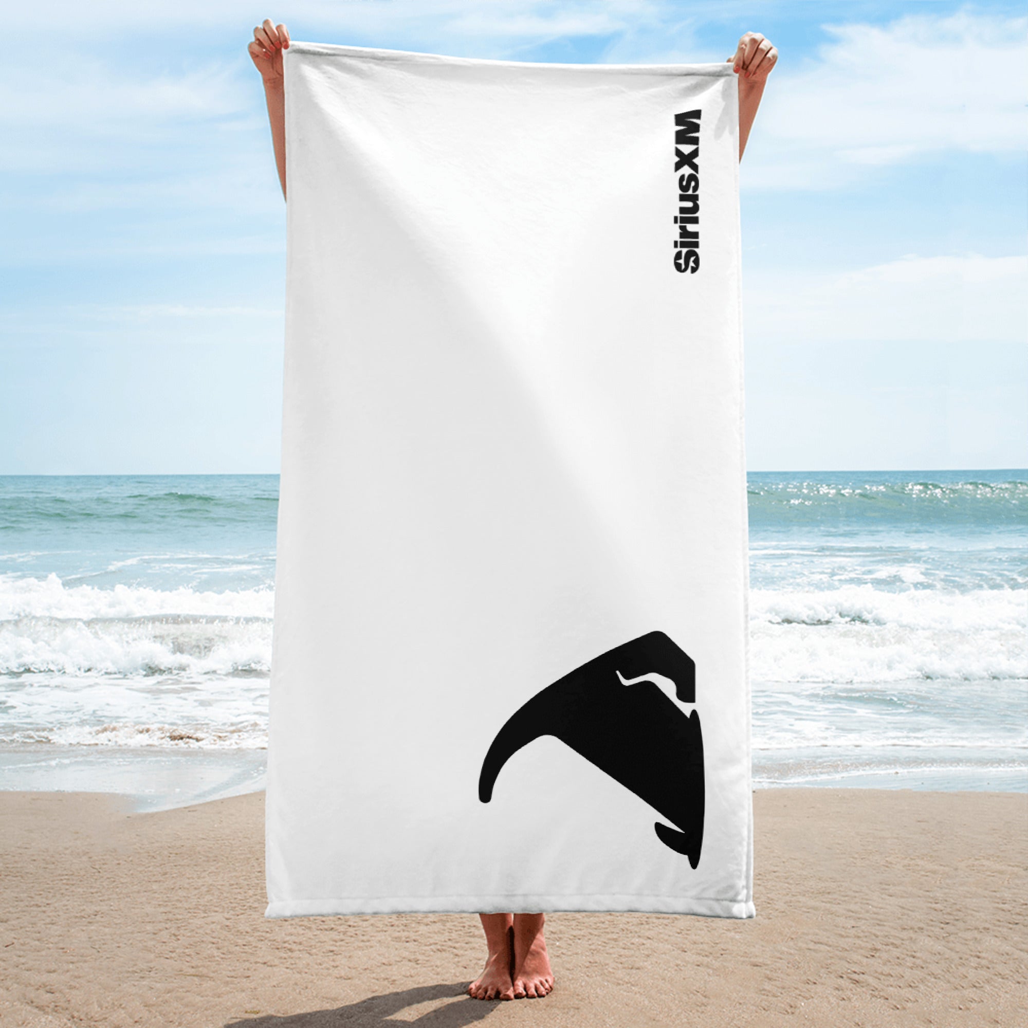 A person stands barefoot on the beach holding a vertically oriented white towel that displays a black silhouette of the back of Stella, the 'SiriusXM' dog mascot, in the bottom right corner. The 'SiriusXM' logo is printed vertically near the top right in black.