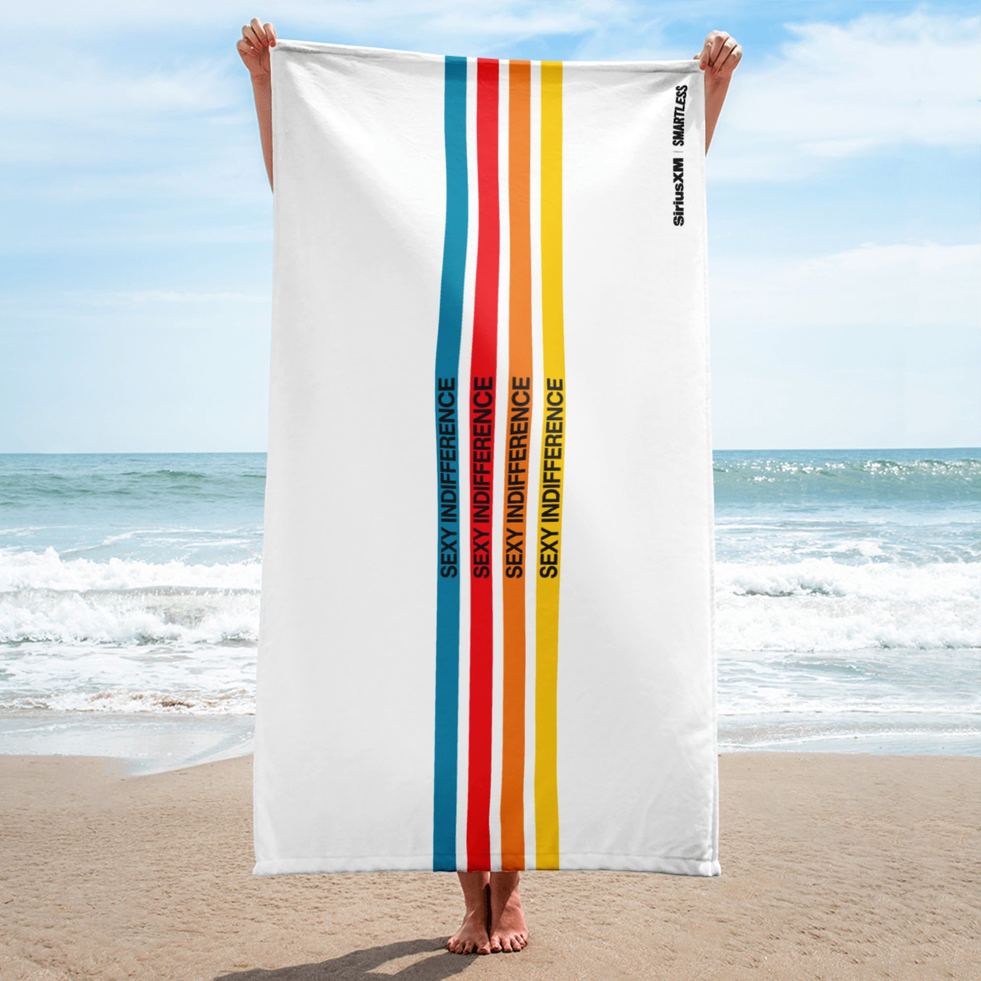 A person stands on the beach holding a vertically oriented white towel with vertical stripes in blue, red, orange, and yellow, each containing the phrase 'SEXY INDIFFERENCE' in black. The 'SiriusXM' and 'SMARTLESS' logos are printed near the top right edge.