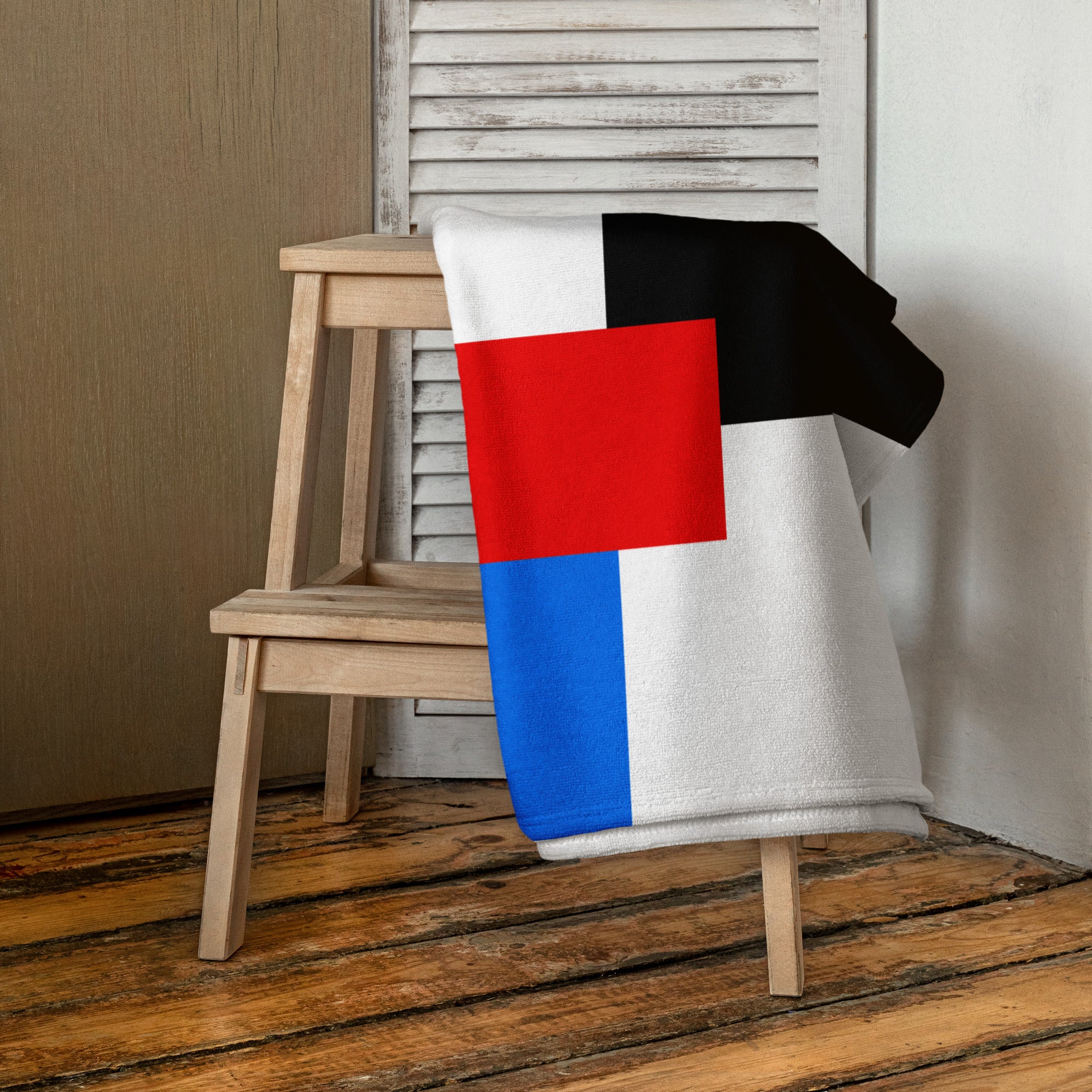 A towel with a modern geometric design in black, red, blue, and white draped over a wooden chair inside a room with a wood floor and white louvered panel wall.