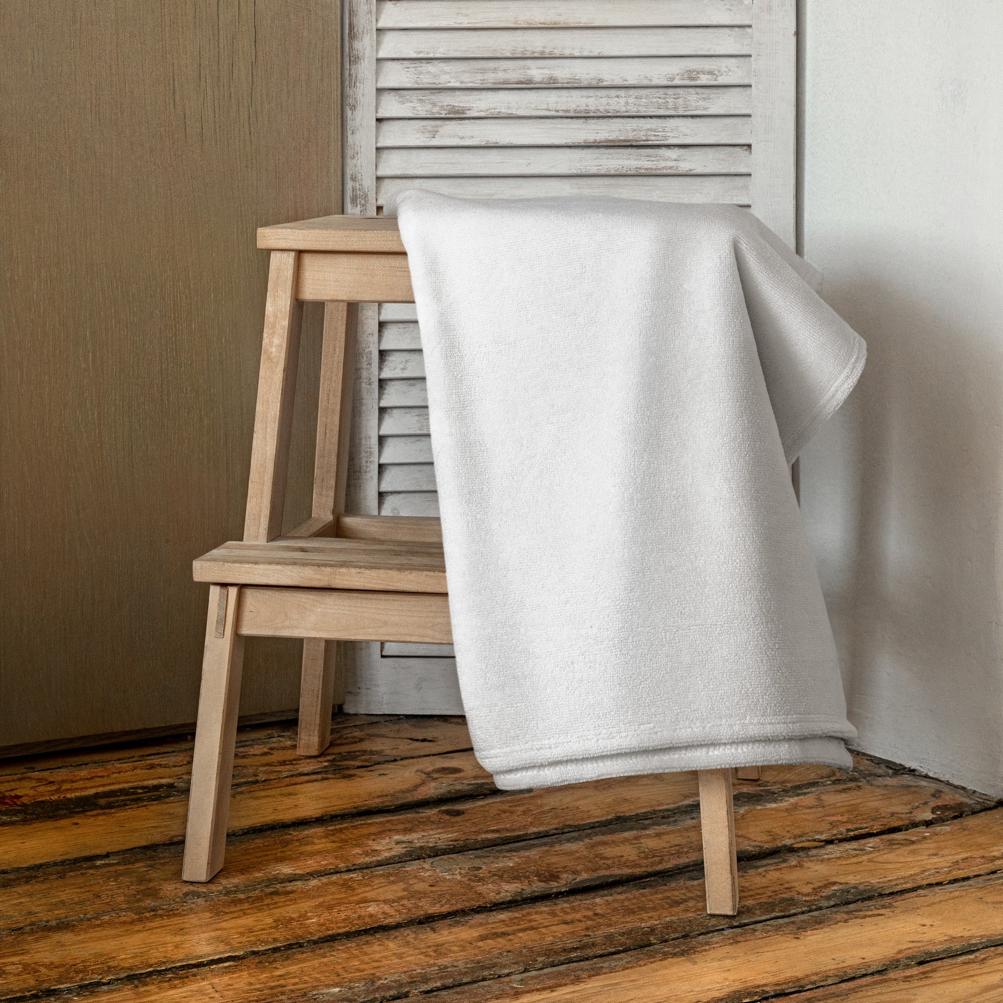 A plain white towel with no visible design, draped over a wooden chair in a rustic indoor setting with a wooden floor and white slatted wall in the background.