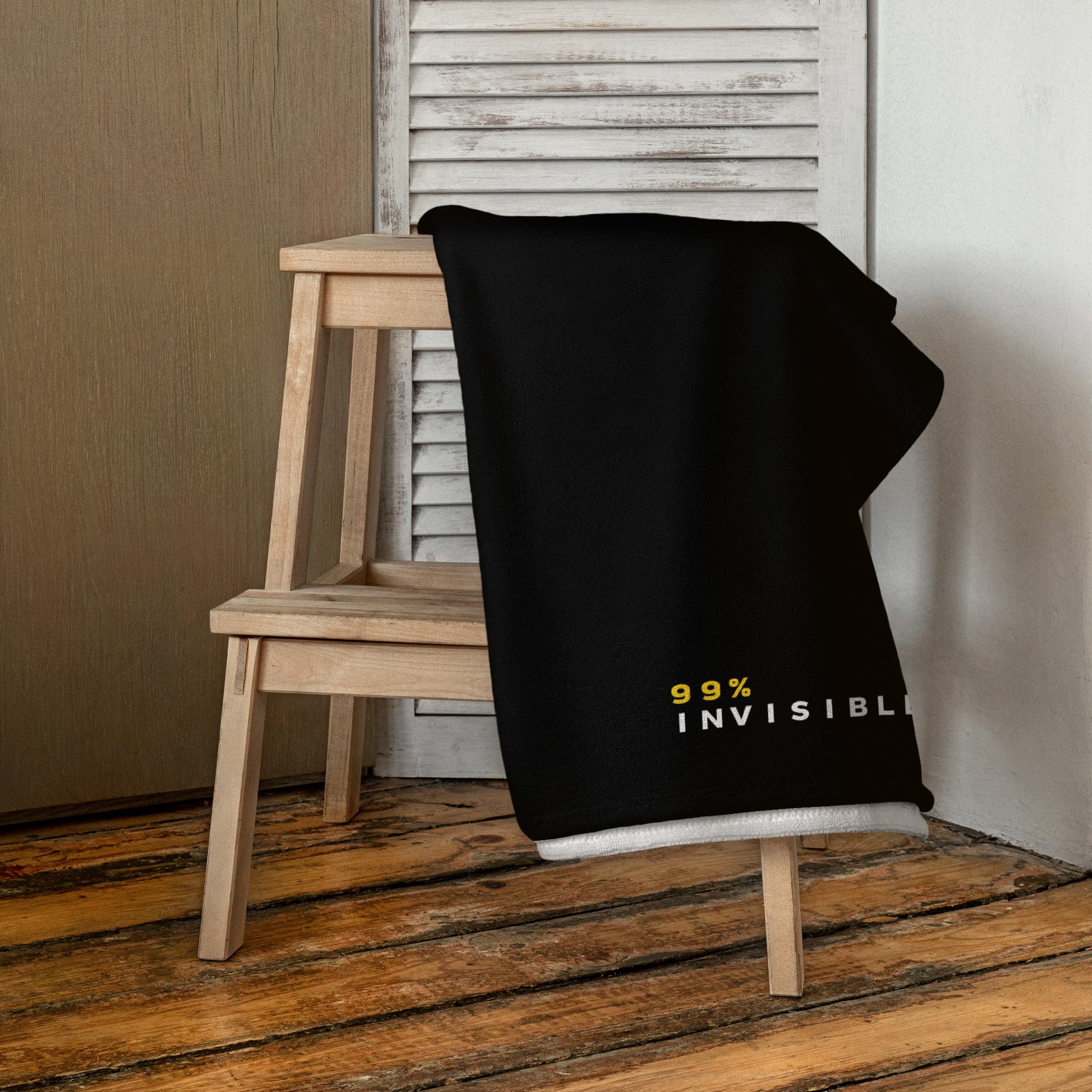 A black towel with '99% INVISIBLE' in white and yellow text draped over a light wooden chair in a cozy room with a wood floor and shutter-style wall.