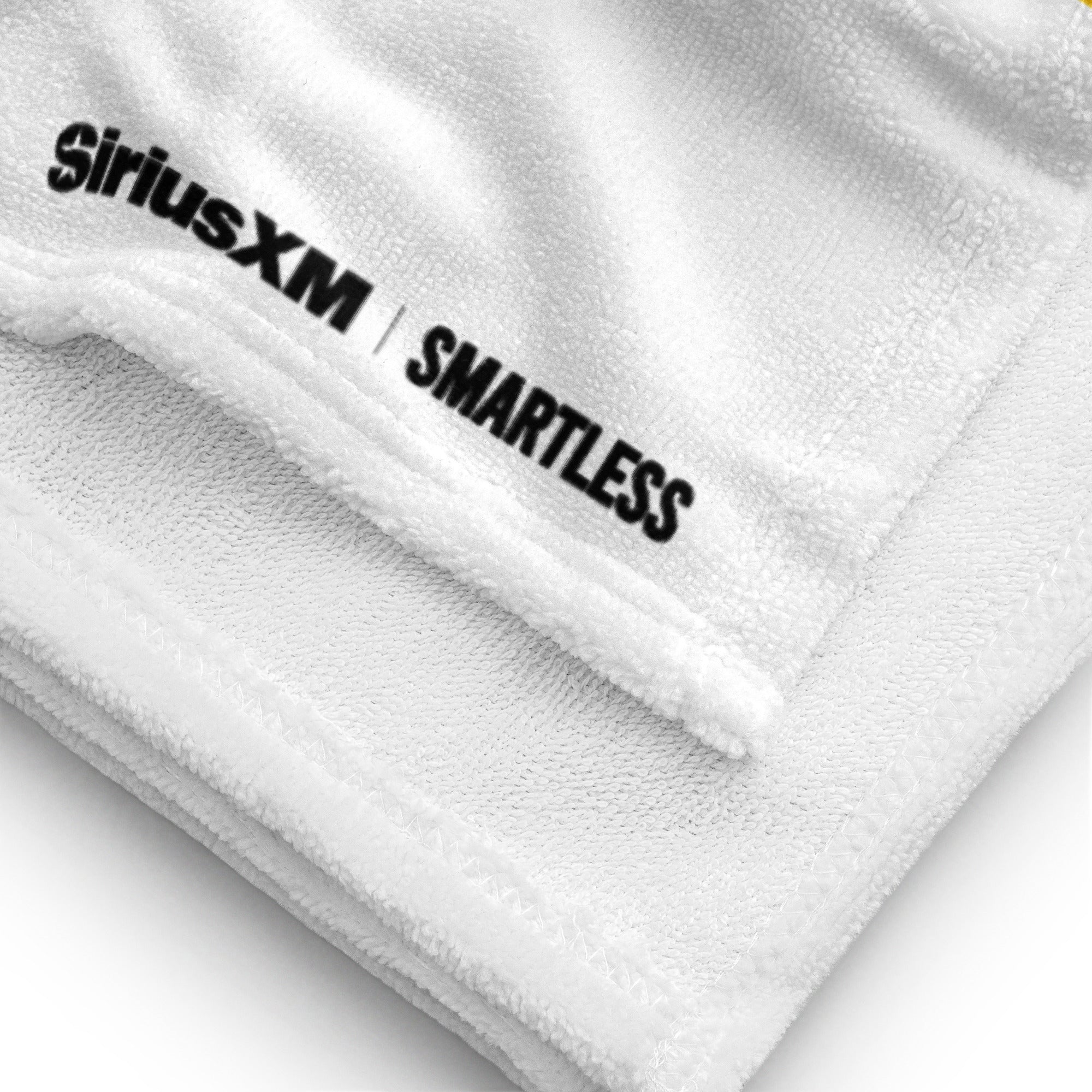 A close-up of the corner of a white towel showing soft terry texture and the 'SiriusXM' and 'SMARTLESS' logos printed in black near the edge seam.