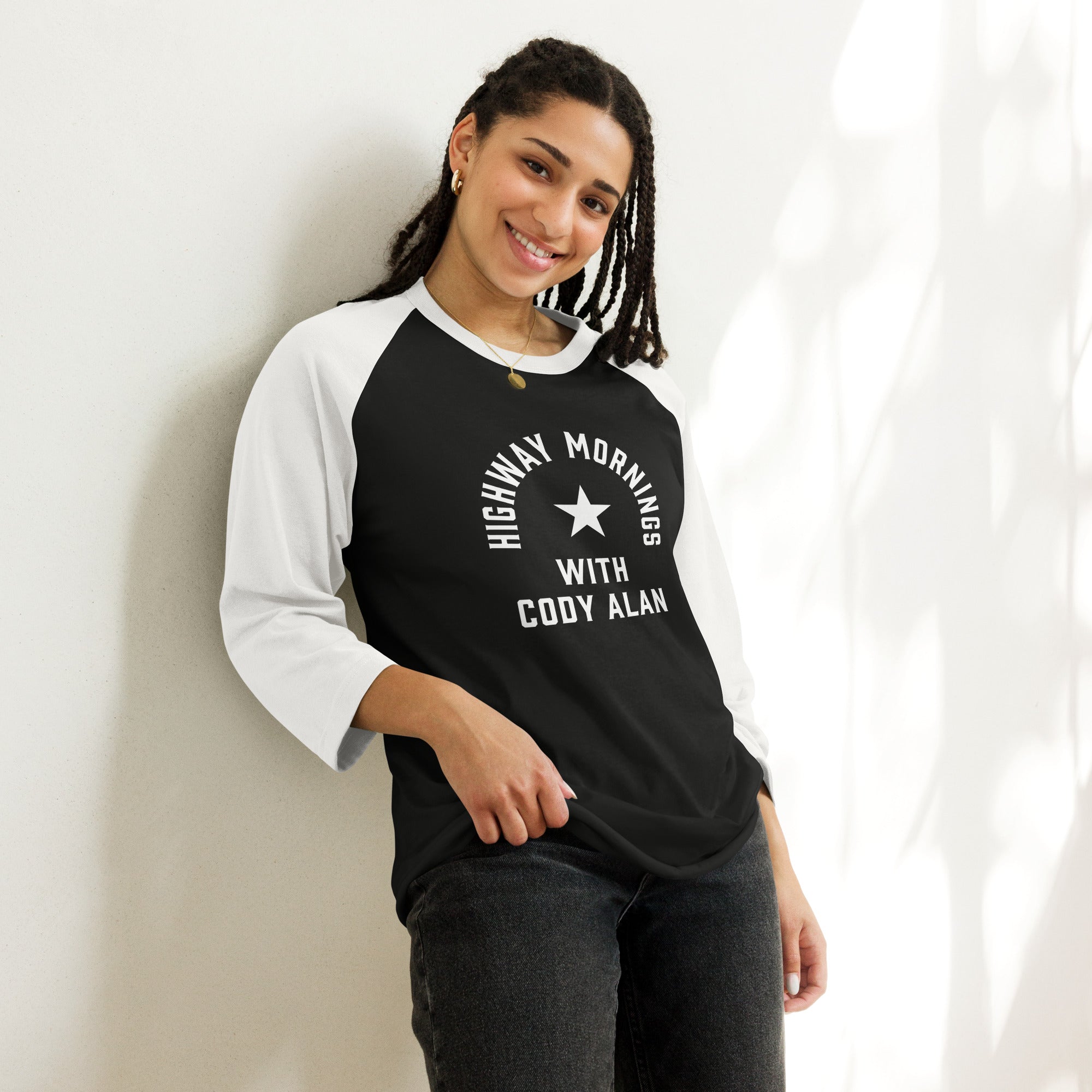 A smiling woman wearing a black and white 3/4-sleeve raglan shirt with the text 'HIGHWAY MORNINGS WITH CODY ALAN' printed in white around a star graphic on the front.