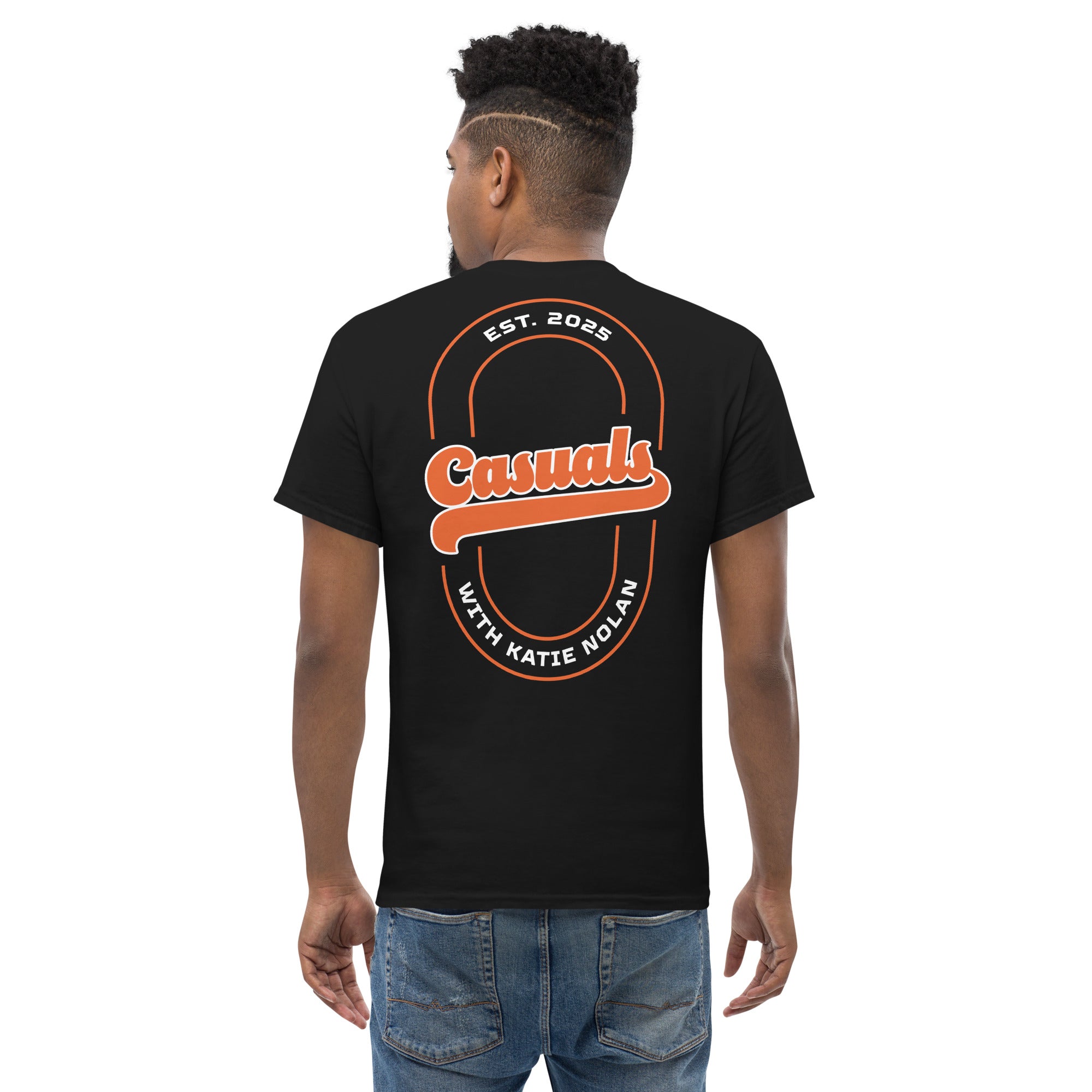 A man shown from behind wearing a black short-sleeve T-shirt, back view. The back features a large orange and white oval graphic with the word 'Casuals' in script across the center, 'EST. 2025' above, and 'WITH KATIE NOLAN' below.