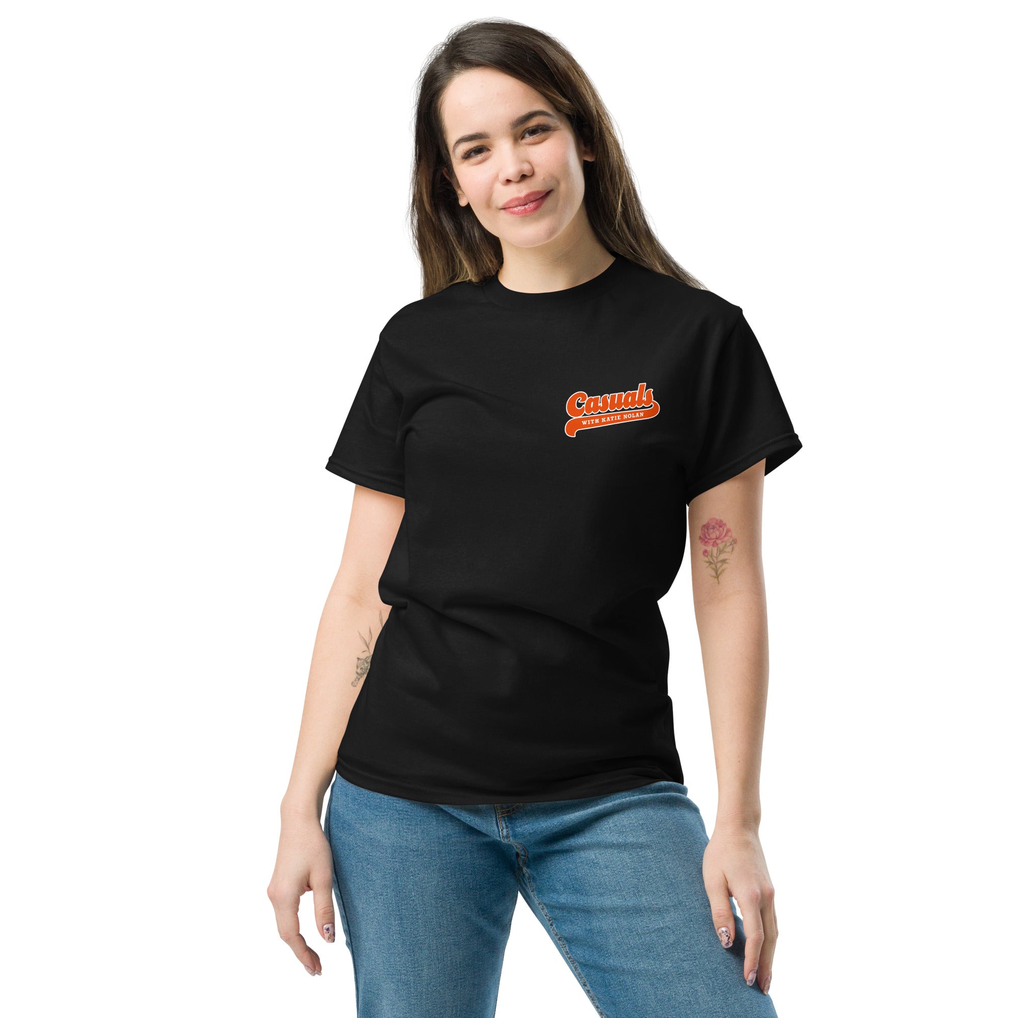 A woman smiling in a front view while wearing a black short-sleeve T-shirt. The front of the shirt features an orange and white logo on the left chest reading 'Casuals WITH KATIE NOLAN.' She stands casually with her hands at her sides. 