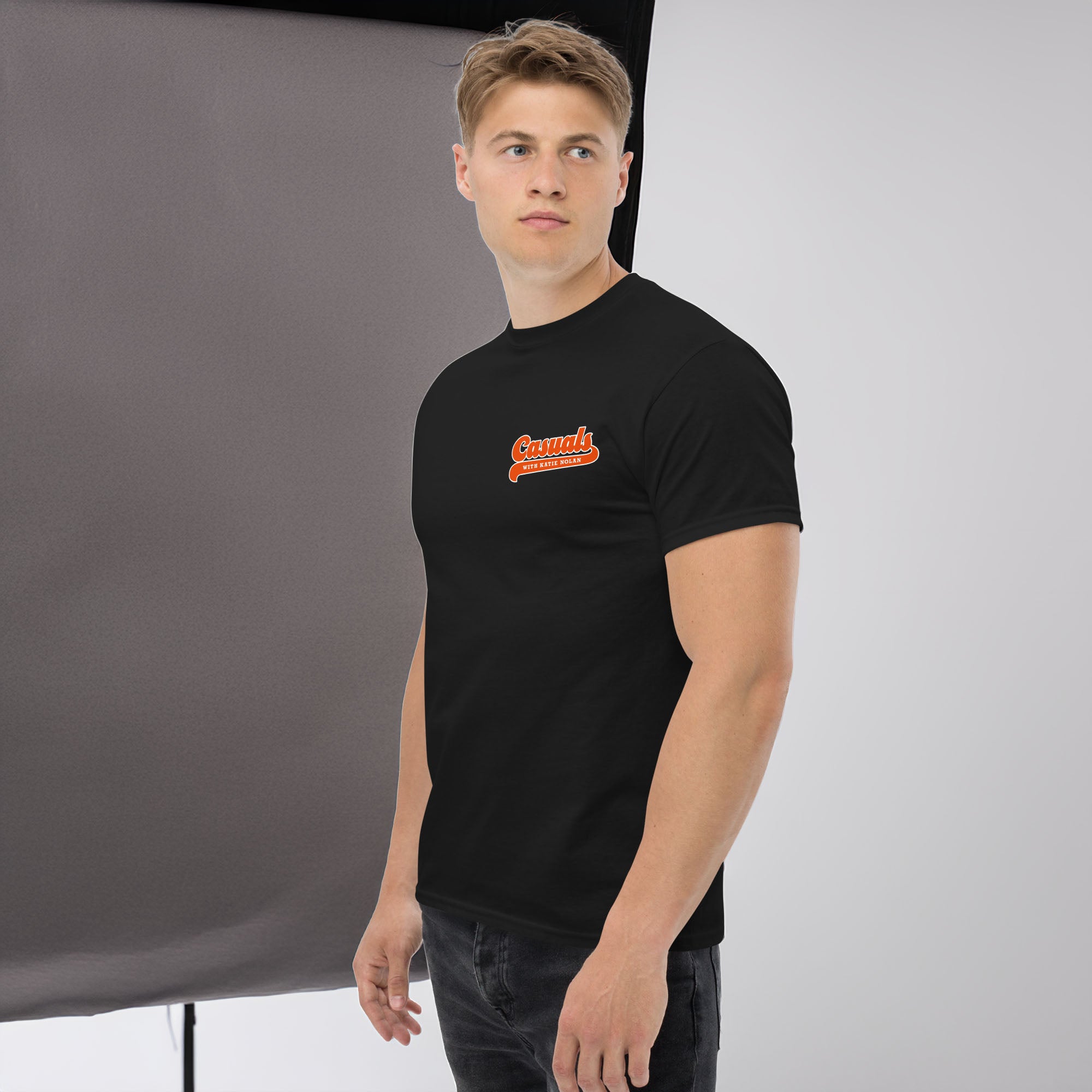 A man facing slightly left wearing a black short-sleeve T-shirt, shown in a left side view. The front design displays an orange and white retro-style logo that reads 'Casuals WITH KATIE NOLAN' on the left chest.