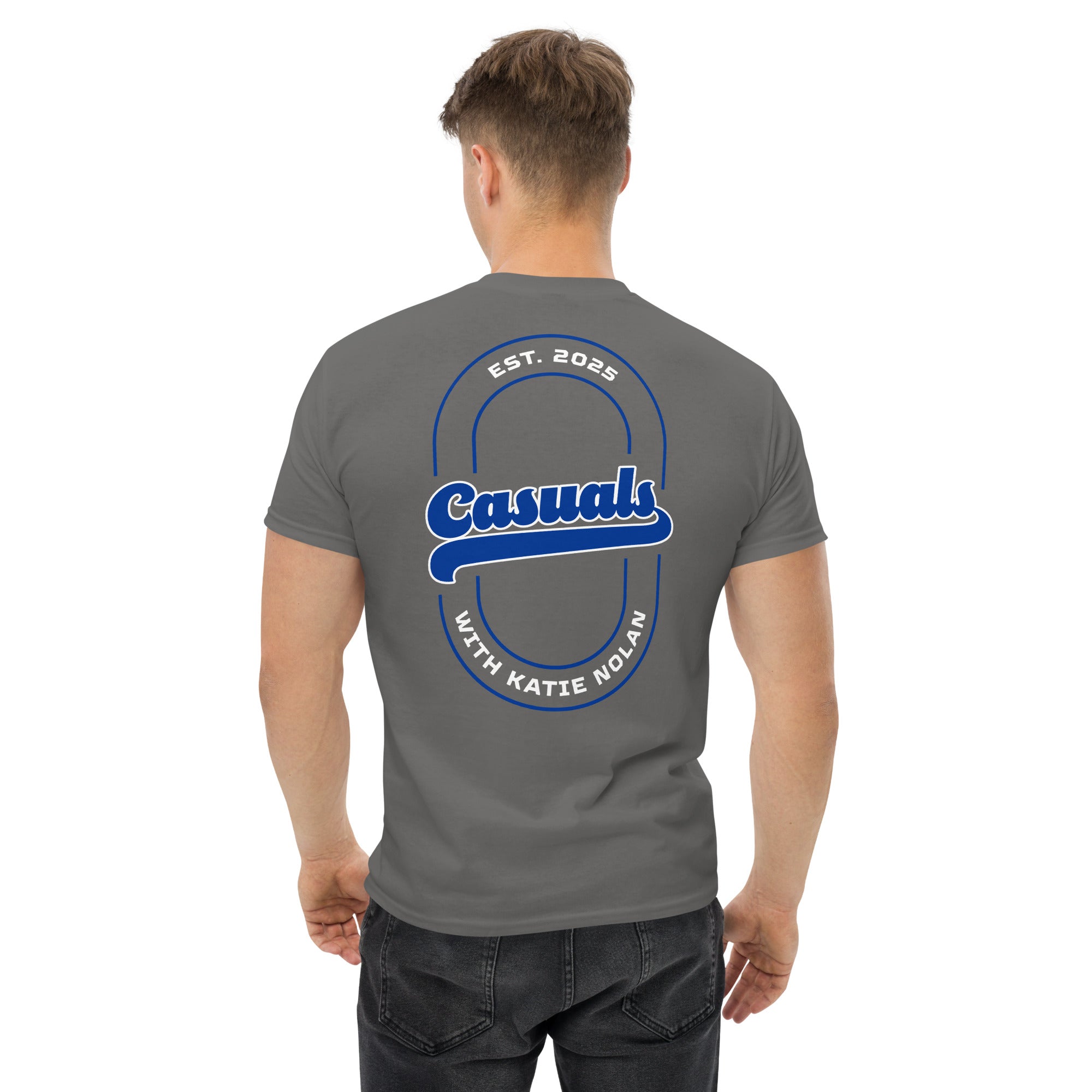 Charcoal short-sleeve t-shirt with a large blue and white back design featuring an oval outline, bold text 'Casuals' in the center, 'EST. 2025' above, and 'WITH KATIE NOLAN' curved below, worn by a man facing away.
