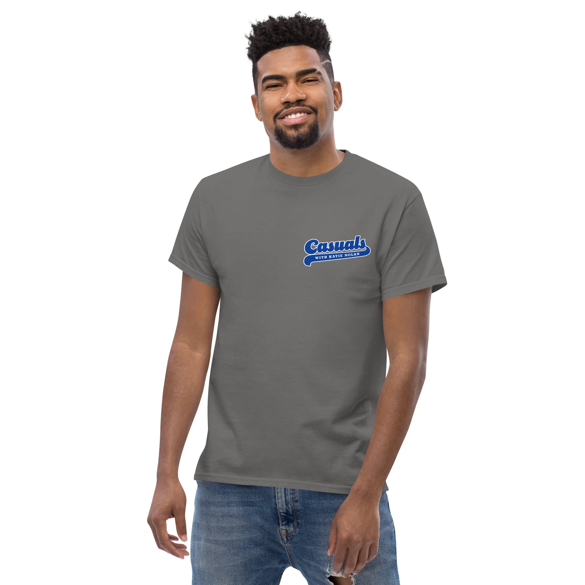 Charcoal short-sleeve t-shirt with a small blue and white left chest logo reading 'Casuals' with 'WITH KATIE NOLAN' underneath, worn by a man standing in jeans, smiling toward the camera.