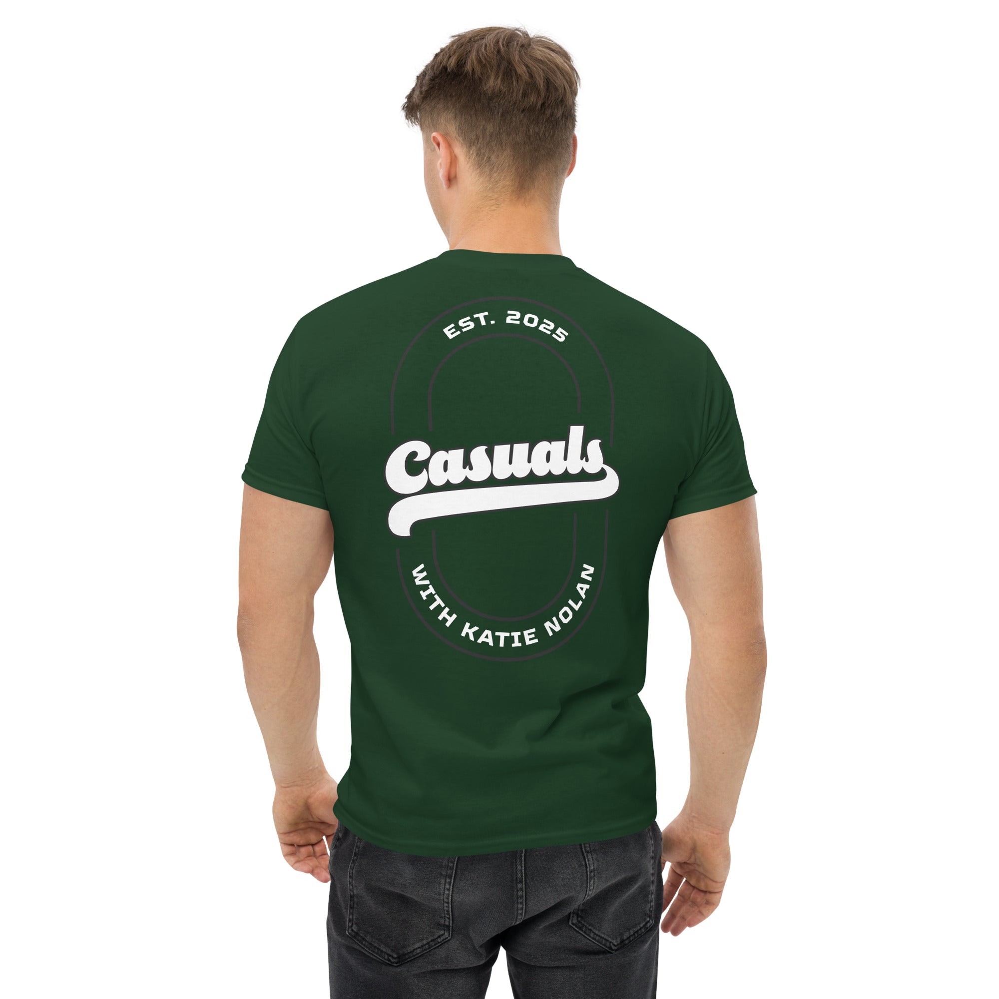 Man wearing a forest green short-sleeve t-shirt with a large white oval outline on the back. Bold white text in the center reads 'Casuals', with 'EST. 2025' above and curved text 'WITH KATIE NOLAN' below.