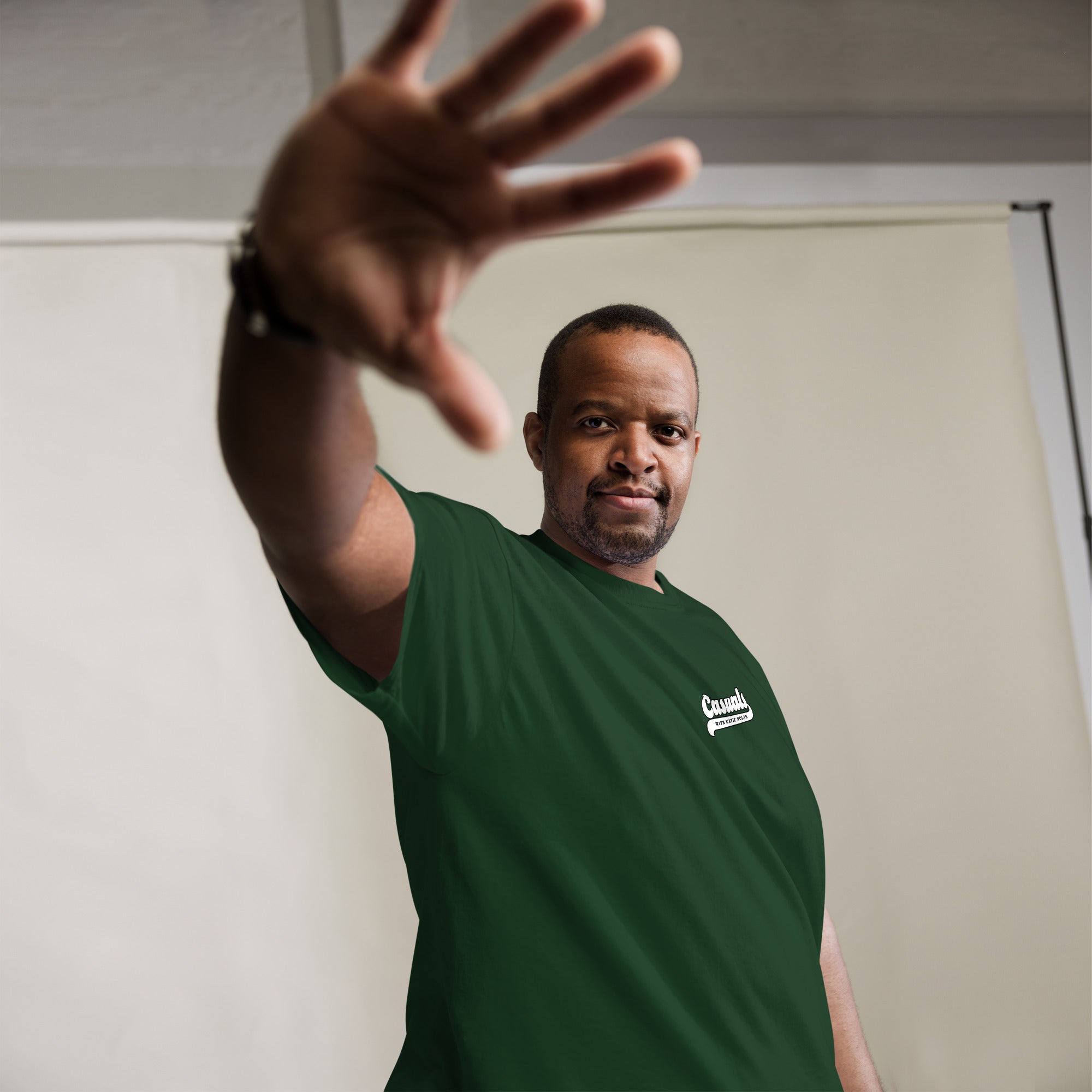 Man wearing a forest green short-sleeve t-shirt with a small white logo on the left chest that reads 'Casuals' with 'WITH KATIE NOLAN' underneath. He is indoors, extending one hand toward the camera in a dynamic pose, standing against a neutral-colored backdrop.