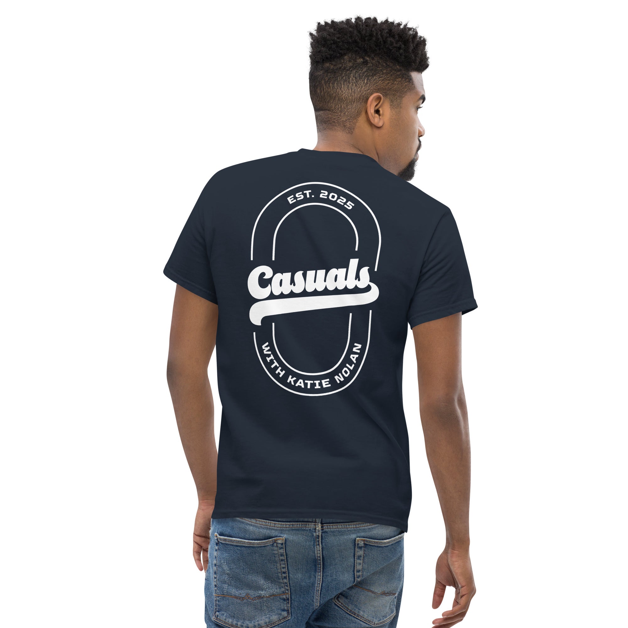 Man wearing a navy short-sleeve t-shirt with a large white back design: oval outline, bold 'Casuals' in the center, 'EST. 2025' above, and 'WITH KATIE NOLAN' curved below.