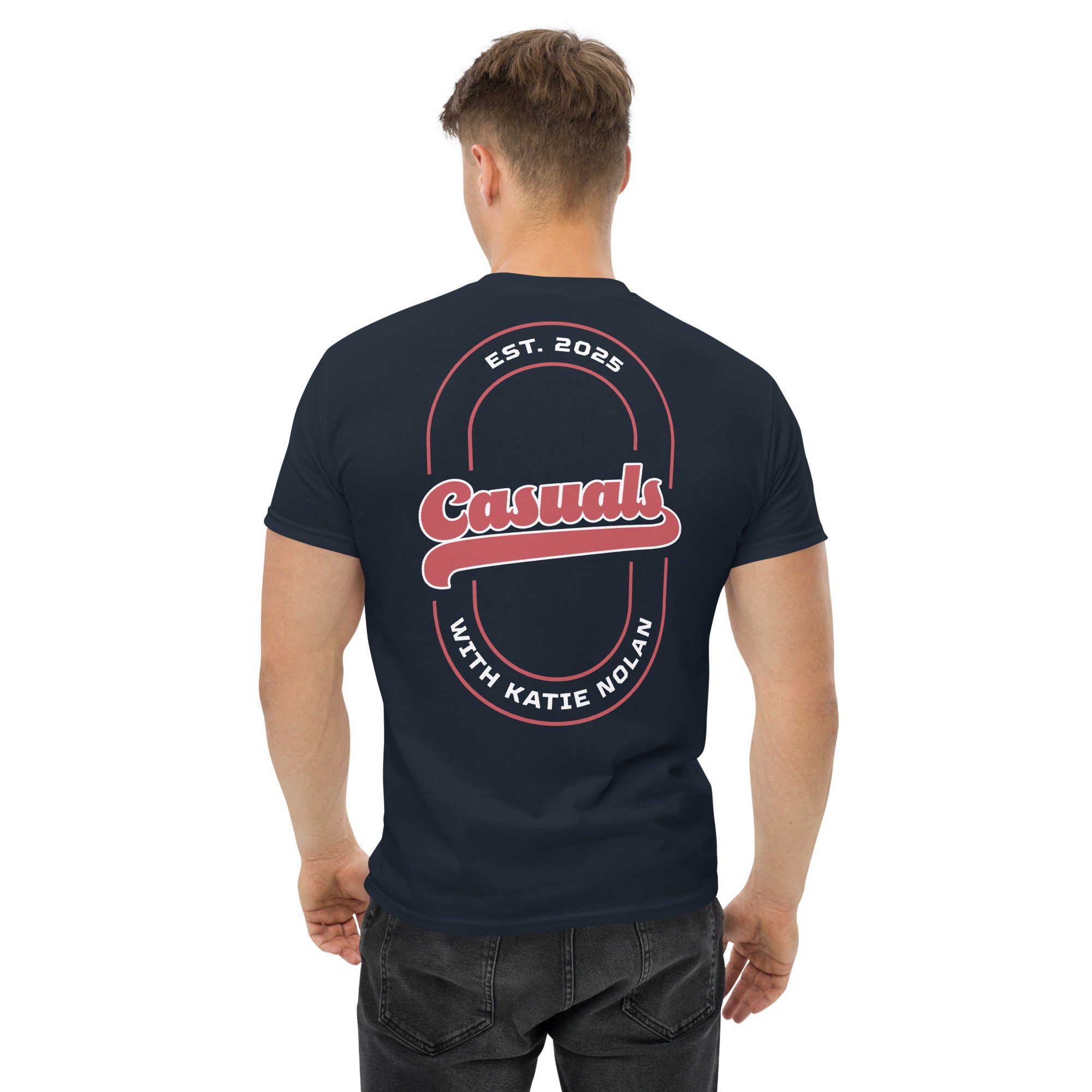 A man shown from the back wearing a navy blue short-sleeve T-shirt. The back of the shirt features a large red and white oval graphic with the word 'Casuals' in script across the center, 'EST. 2025' above, and 'WITH KATIE NOLAN' below.