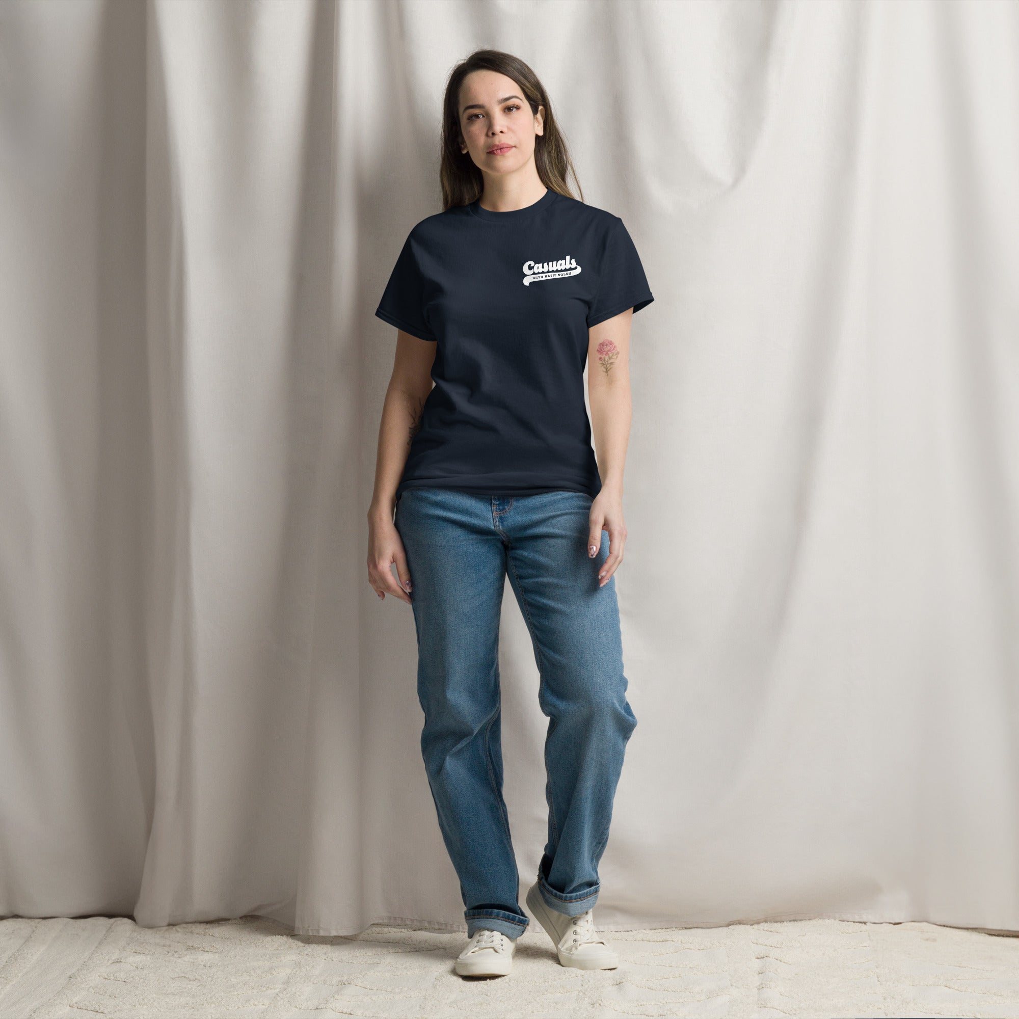 Woman wearing a navy short-sleeve t-shirt with a small white chest logo on the left that reads 'Casuals' with 'WITH KATIE NOLAN' below. She is standing on a carpeted floor against a backdrop of soft beige curtains, wearing light blue jeans and white sneakers.