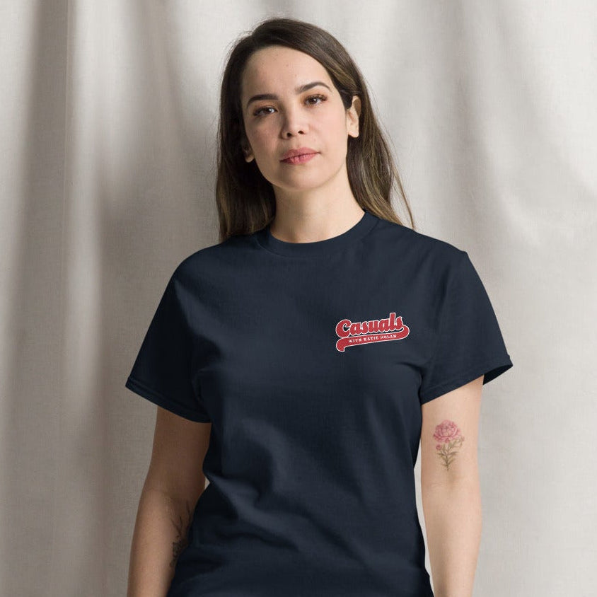 A woman shown from the front wearing a navy blue short-sleeve T-shirt. The front of the shirt features a small red and white logo on the left chest that reads 'Casuals WITH KATIE NOLAN.'