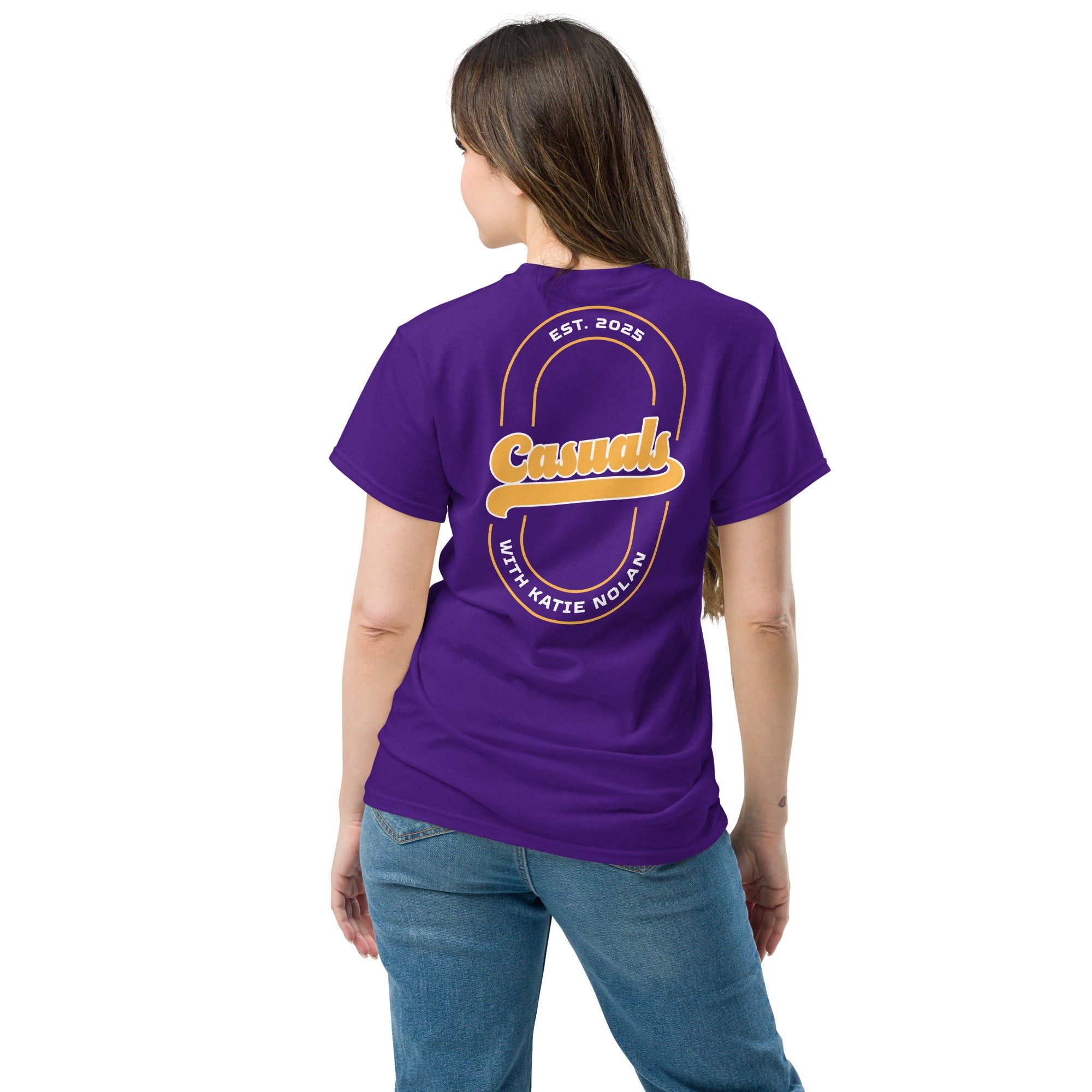 A woman shown from behind wearing a purple short-sleeve T-shirt with a large back design. The graphic features the word 'Casuals' in bold yellow lettering outlined in white, centered within a double-line oval with the text 'EST. 2025' at the top and 'WITH KATIE NOLAN' at the bottom.