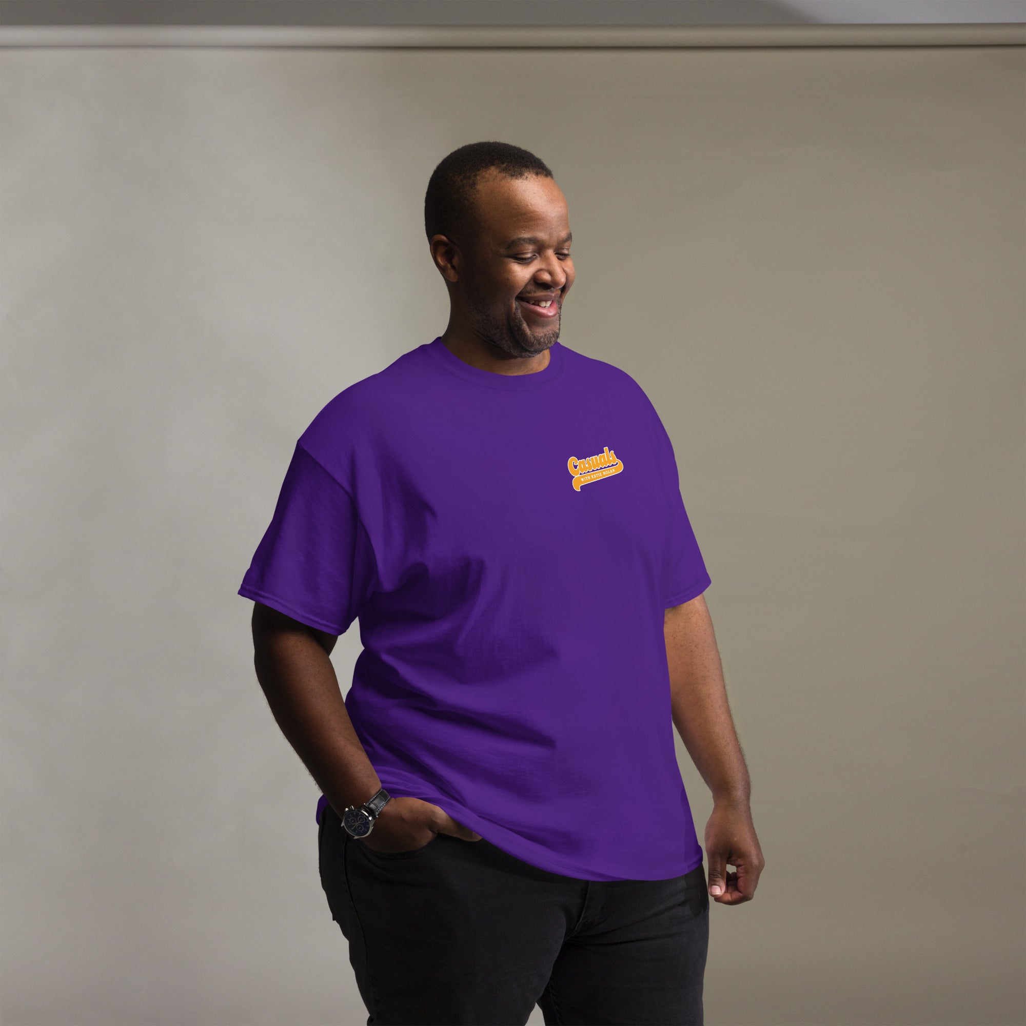 man smiling and looking down while wearing a purple short-sleeve T-shirt. The left chest shows a small yellow and white logo that reads 'Casuals WITH KATIE NOLAN' in retro-style text.