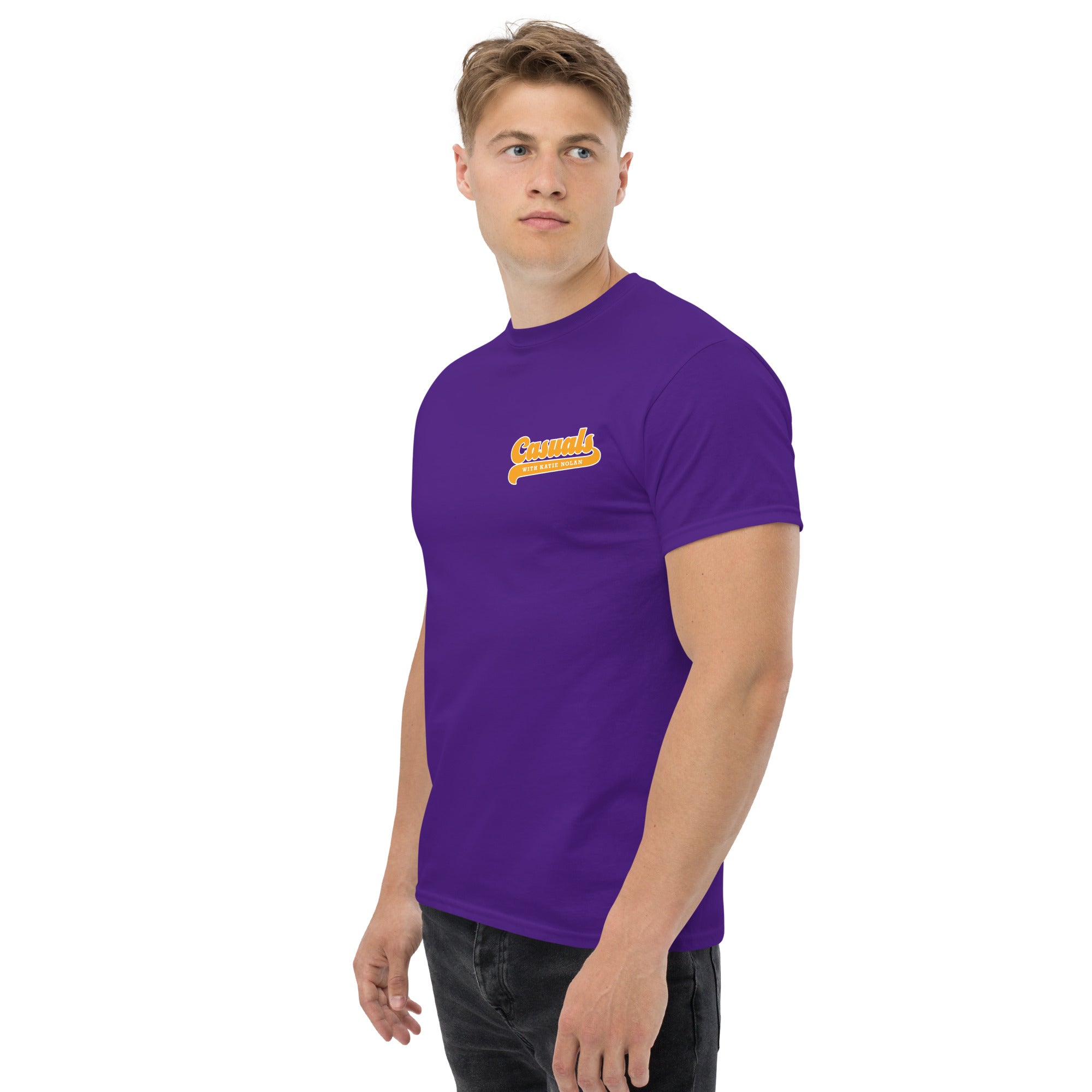 A man facing slightly to the left while wearing a bright purple short-sleeve T-shirt. A small yellow and white logo on the left chest reads 'Casuals WITH KATIE NOLAN' in retro script.
