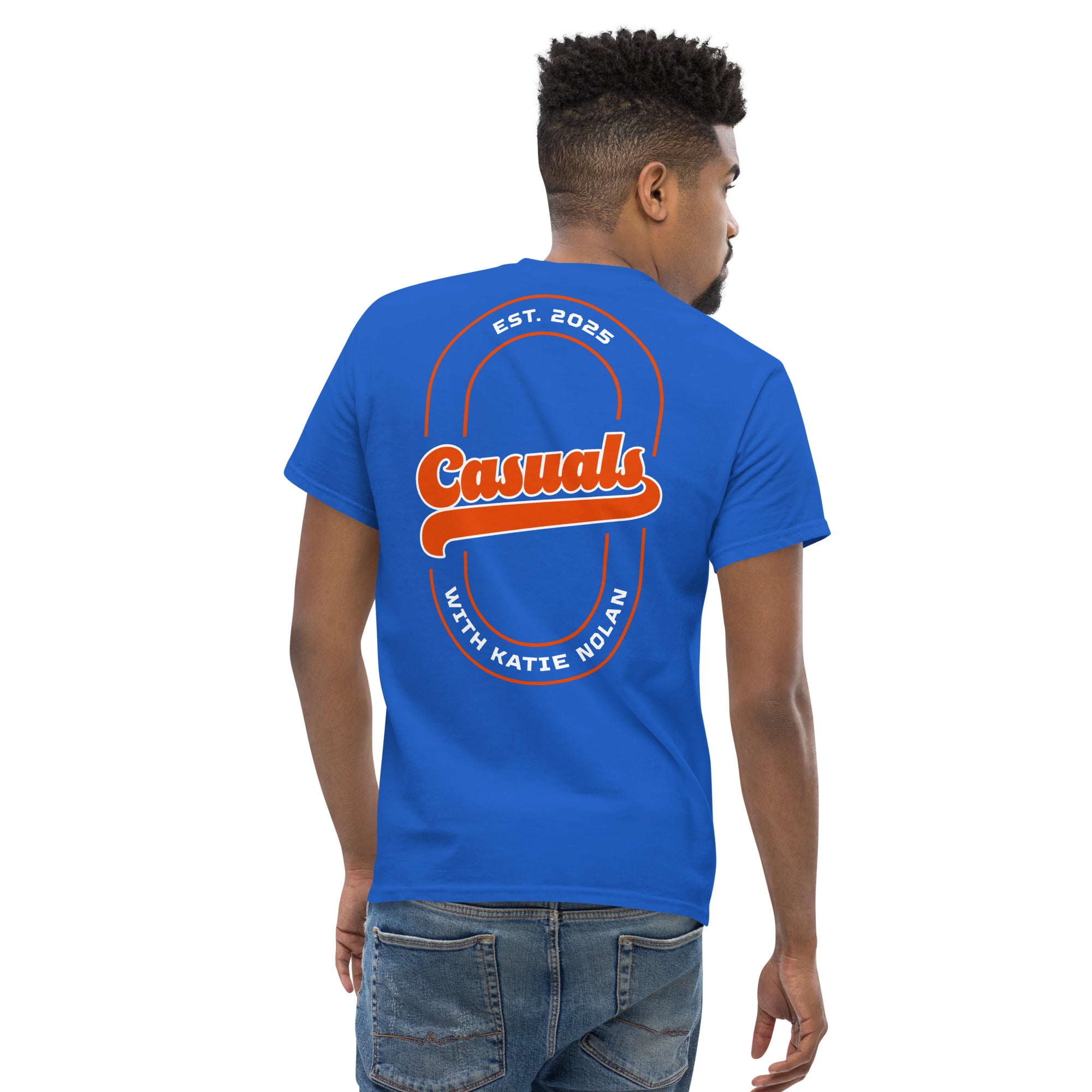 A man shown from the back wearing a bright royal blue short-sleeve T-shirt. The back design features large orange and white text that reads 'Casuals' in a retro script, with 'EST. 2025' above and 'WITH KATIE NOLAN' below, enclosed in a double-line oval outline.