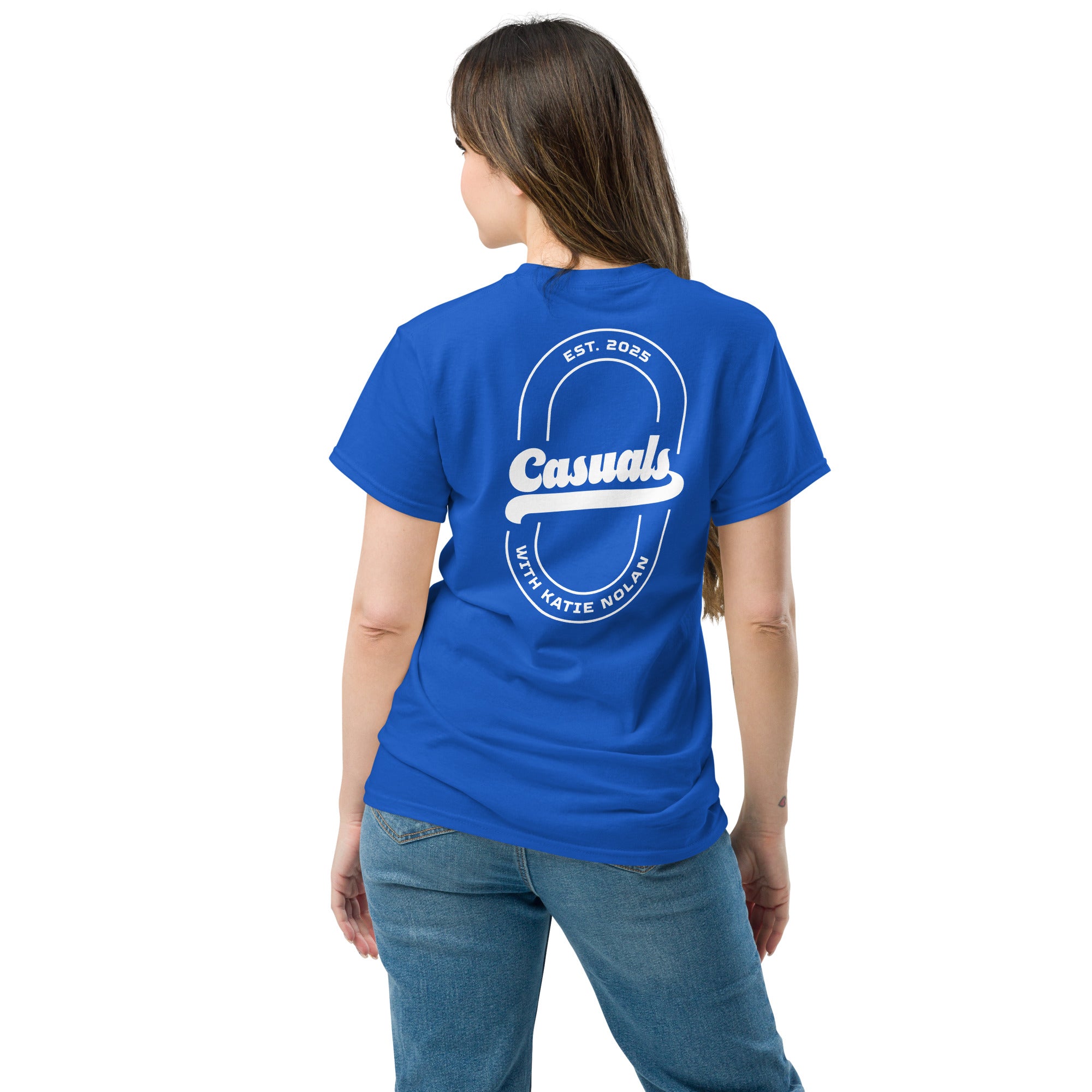 Woman wearing a royal blue short-sleeve t-shirt with a large white oval outline on the back. Inside, bold white text reads 'Casuals' with 'EST. 2025' above and curved text 'WITH KATIE NOLAN' below.