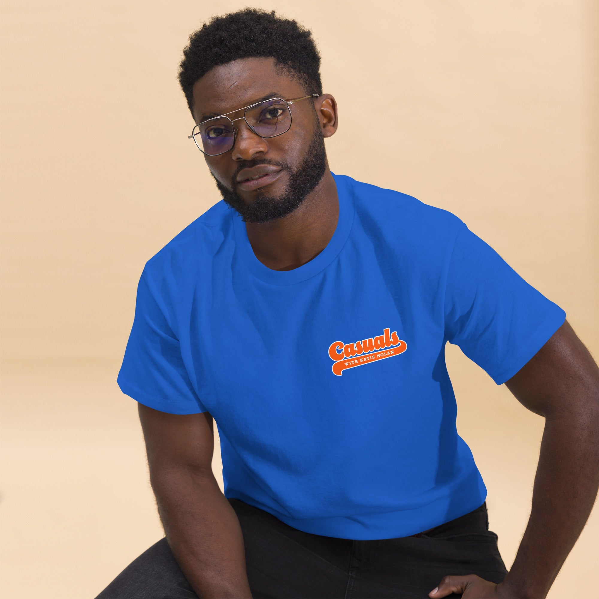 A man sitting casually and looking at the camera while wearing a royal blue T-shirt. A small orange and white front logo reads 'Casuals WITH KATIE NOLAN' in retro script.