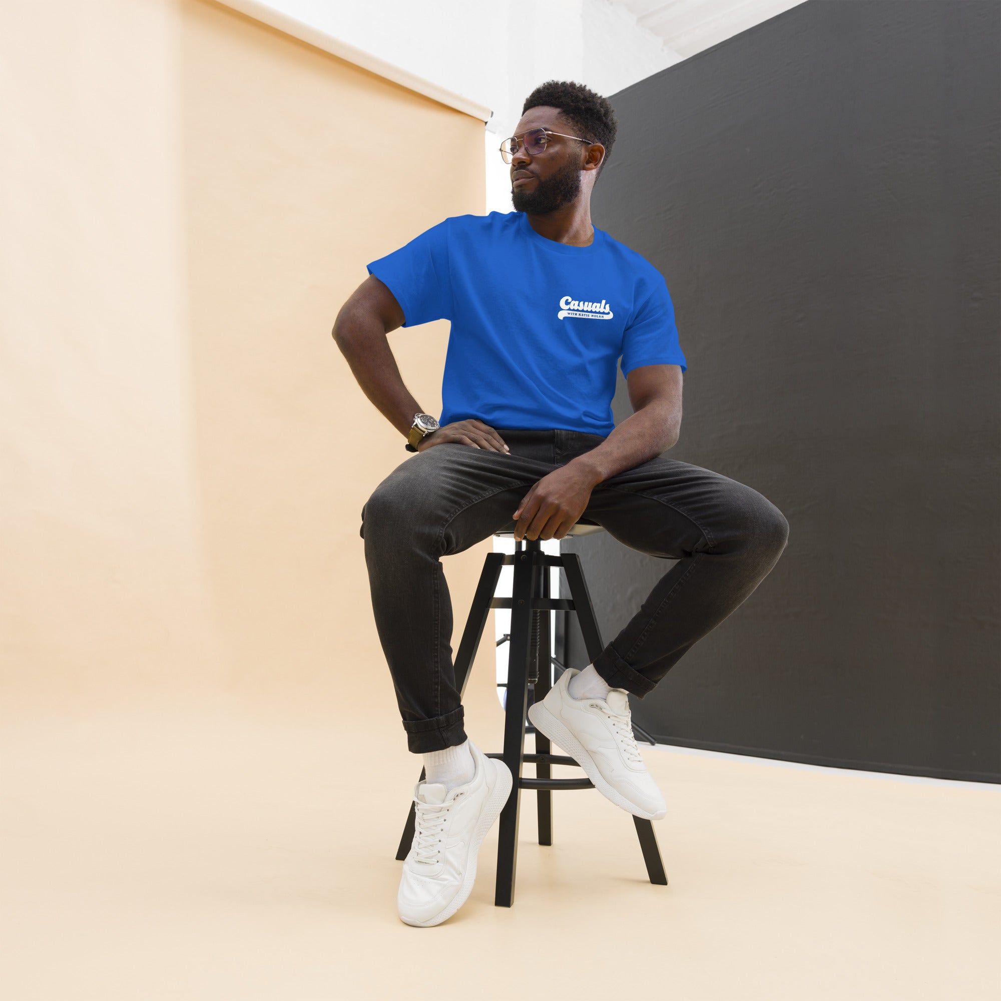 Man wearing a royal blue short-sleeve t-shirt with a small white chest logo on the left reading 'Casuals' with 'WITH KATIE NOLAN' underneath. He is seated on a tall black stool in a photo studio with a beige backdrop and black panel wall, dressed in dark jeans and white sneakers.