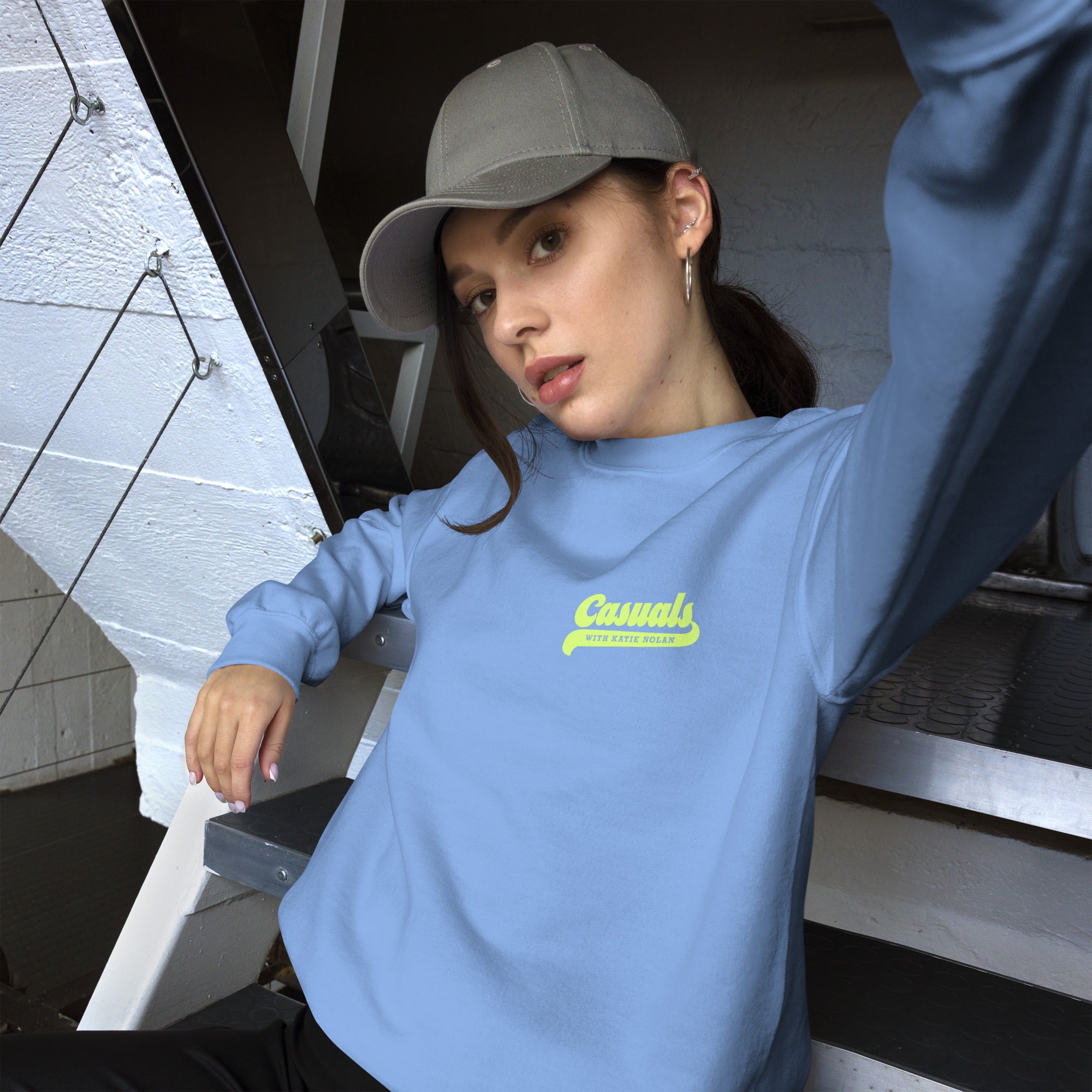 Woman wearing a gray baseball cap, leaning back on stairs while wearing the light blue sweatshirt with 'Casuals' in large yellow script and 'WITH KATIE NOLAN' in smaller yellow text on the front left chest.