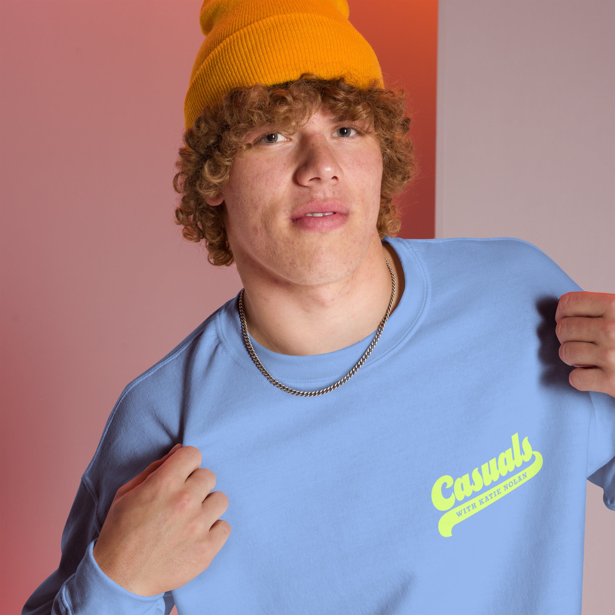 Close-up of person pointing to the left chest design of the sweatshirt, which features the yellow text 'Casuals' in script and 'WITH KATIE NOLAN' below it.