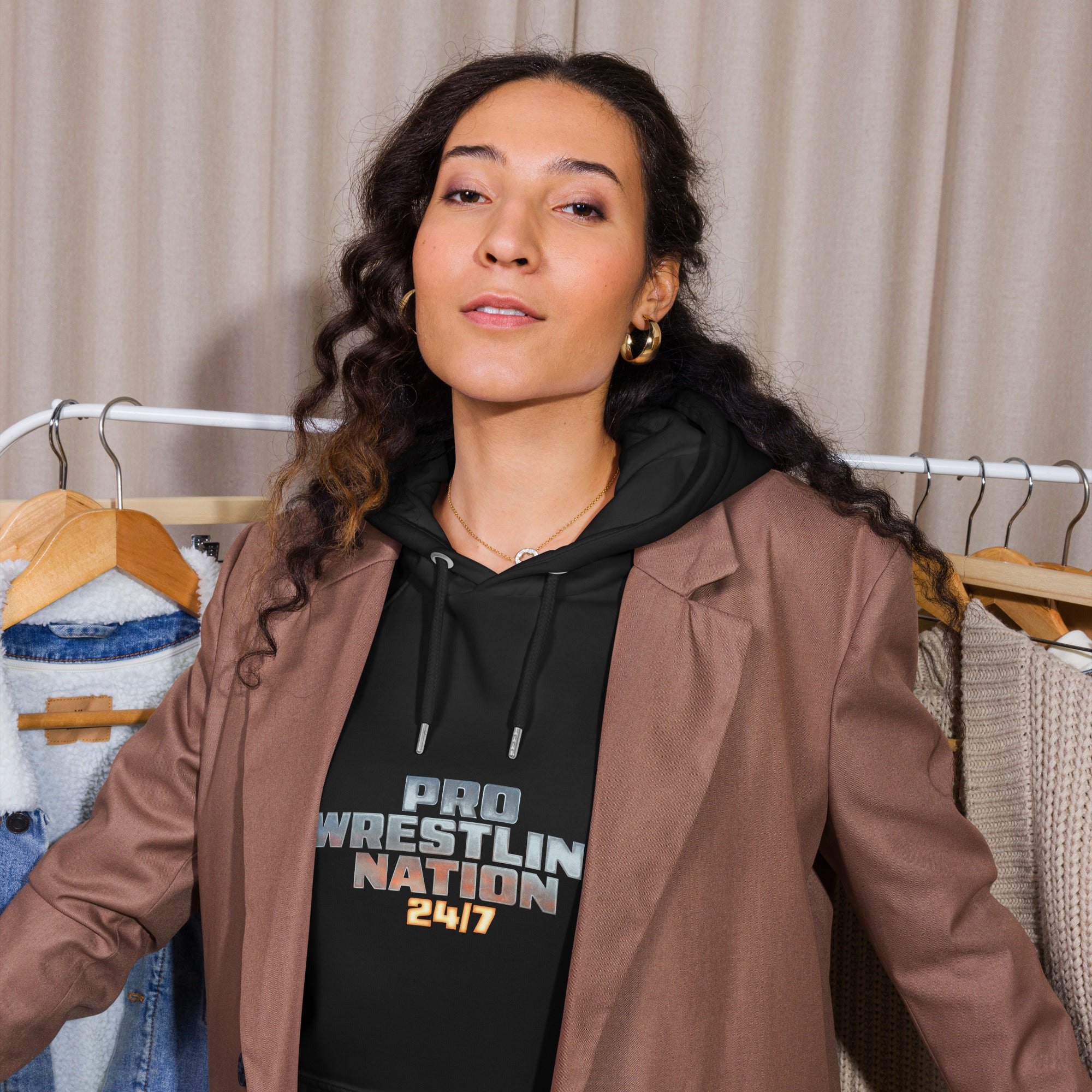 Lifestyle image of a woman wearing the 'Pro Wrestling Nation 24/7' hoodie under a brown blazer, standing in front of a clothing rack.